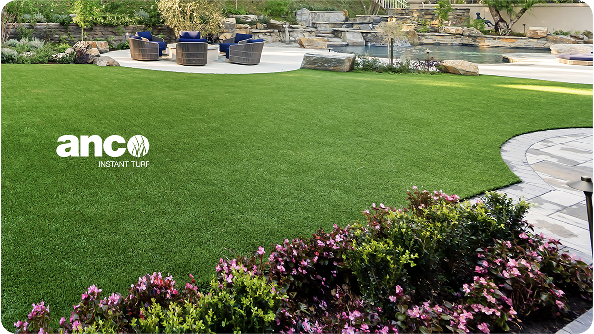 A backyard with well-maintained artificial green turf, a flower bed with pink blossoms, a stone patio, and a pool with a seating area in the background.