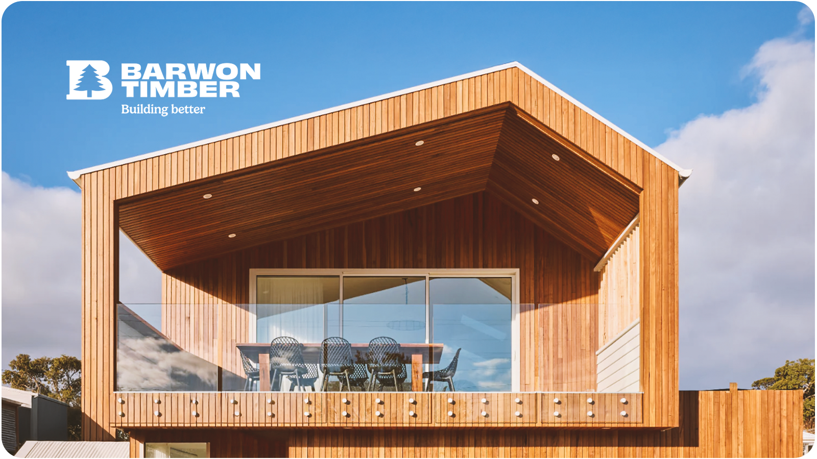 Modern wooden house with a balcony, glass railing, outdoor dining table, and chairs against a blue sky with clouds.