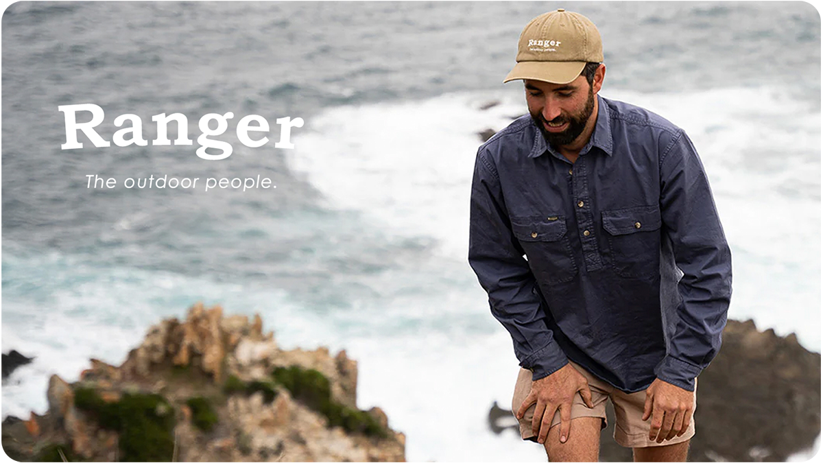 A man smiling near the ocean, wearing a beige cap and navy blue outdoor shirt, leaning down slightly.