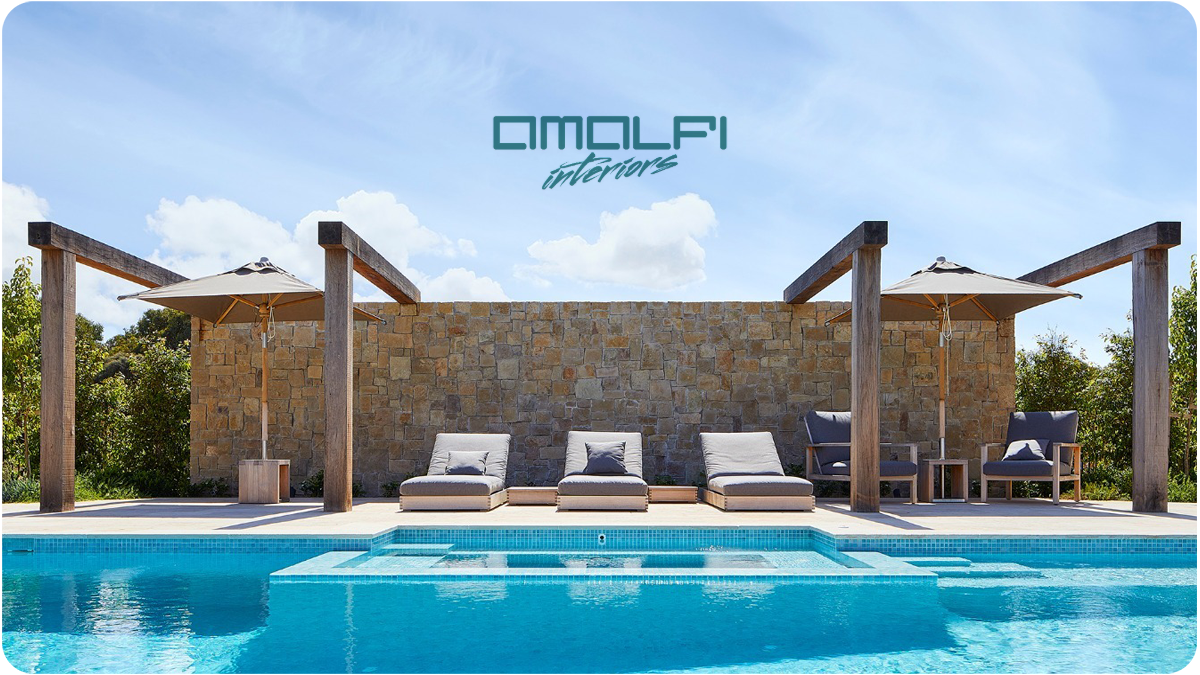 A luxurious outdoor pool area with lounge chairs, umbrellas, a stone wall, and a blue sky with clouds, featuring the text 'AMALFI interiors' at the top.