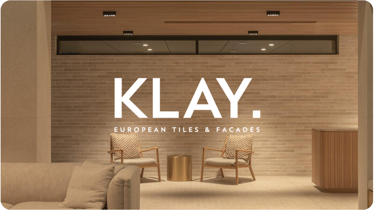 Interior of a modern lobby with two armchairs, a small gold side table, a beige sofa with pillows, and a brick wall with large illuminated sign reading 'KLAY. European Tiles & Facades.' ceiling lights and a large window or reflection above.