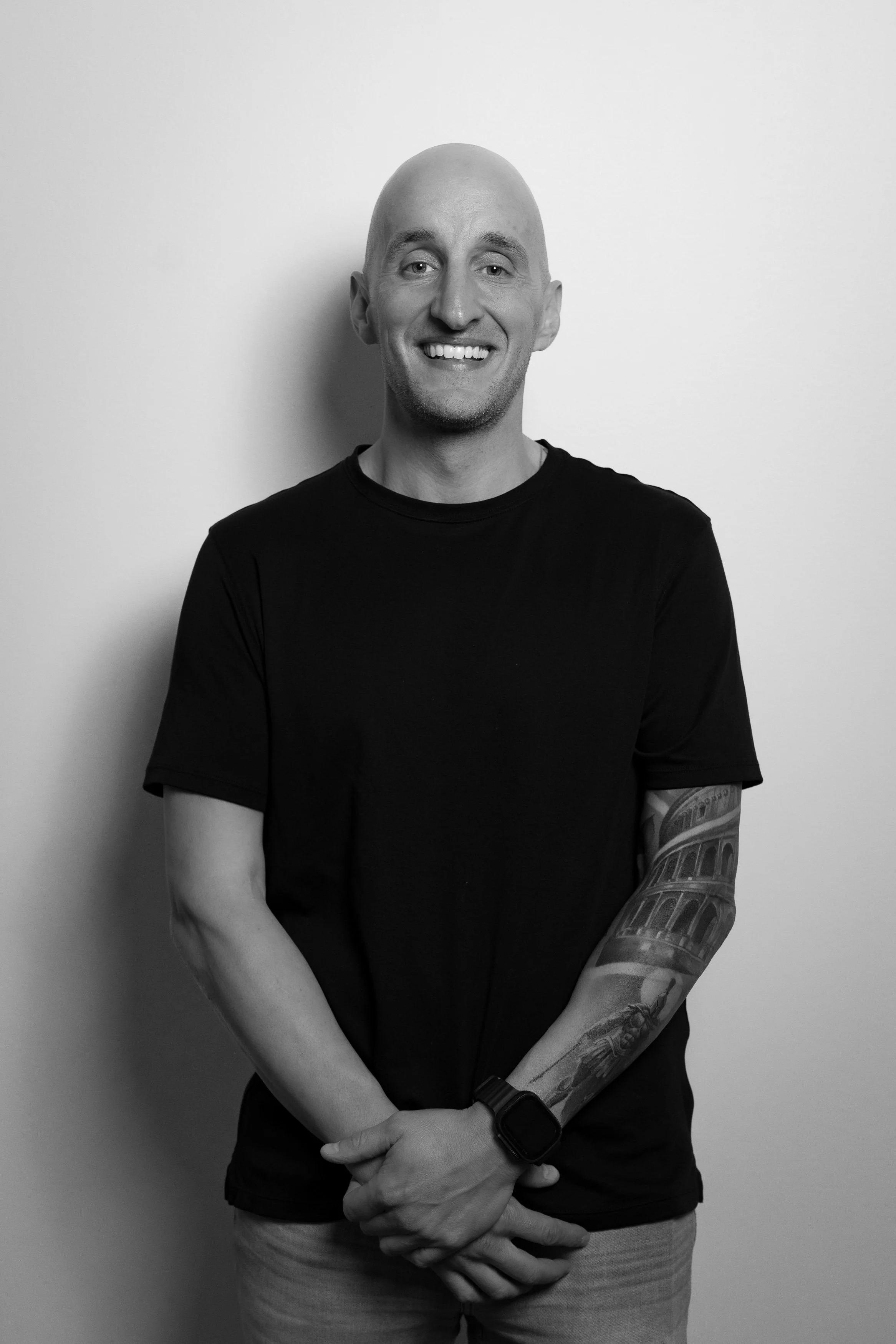 Black and white portrait of a smiling man with a shaved head, wearing a black t-shirt, and displaying a tattoo sleeve on his right arm with a cityscape and structures.