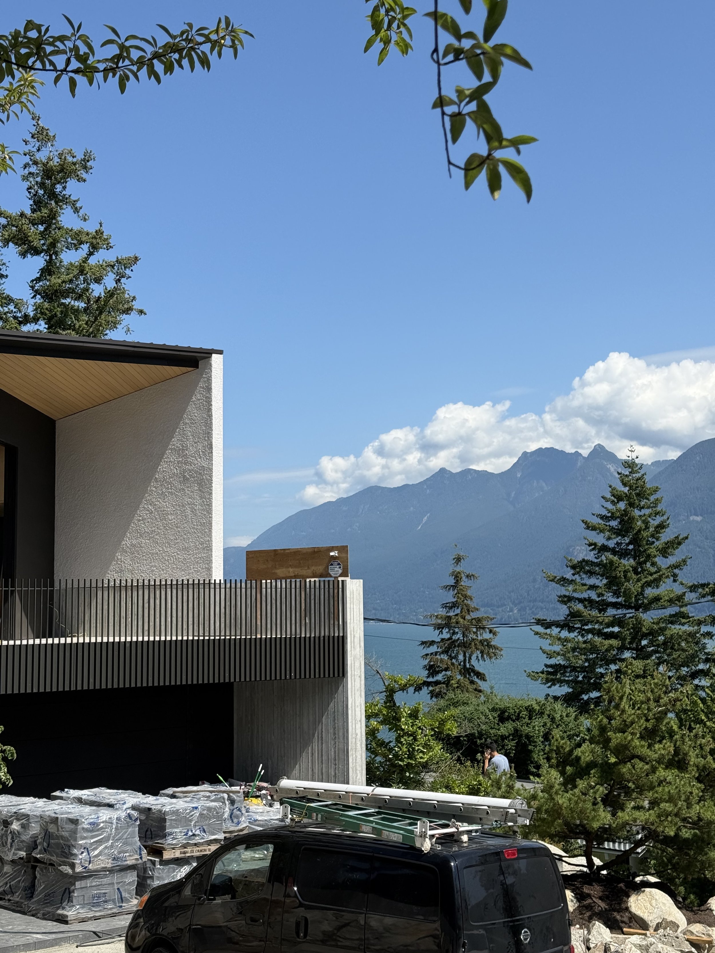 This image shows a modern house with a mountain range in the background. In the foreground, there are construction materials and tools, a ladder on top of a black vehicle, and some people working among green trees.