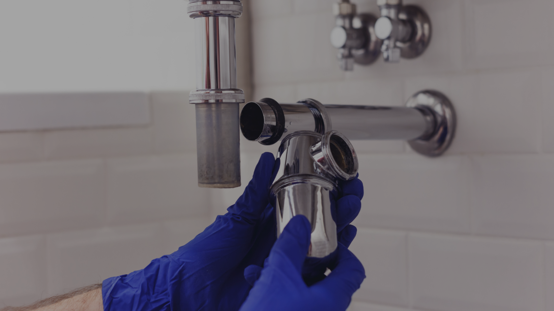 A person wearing blue gloves is inspecting or installing a chrome plumbing pipe under a bathroom sink.