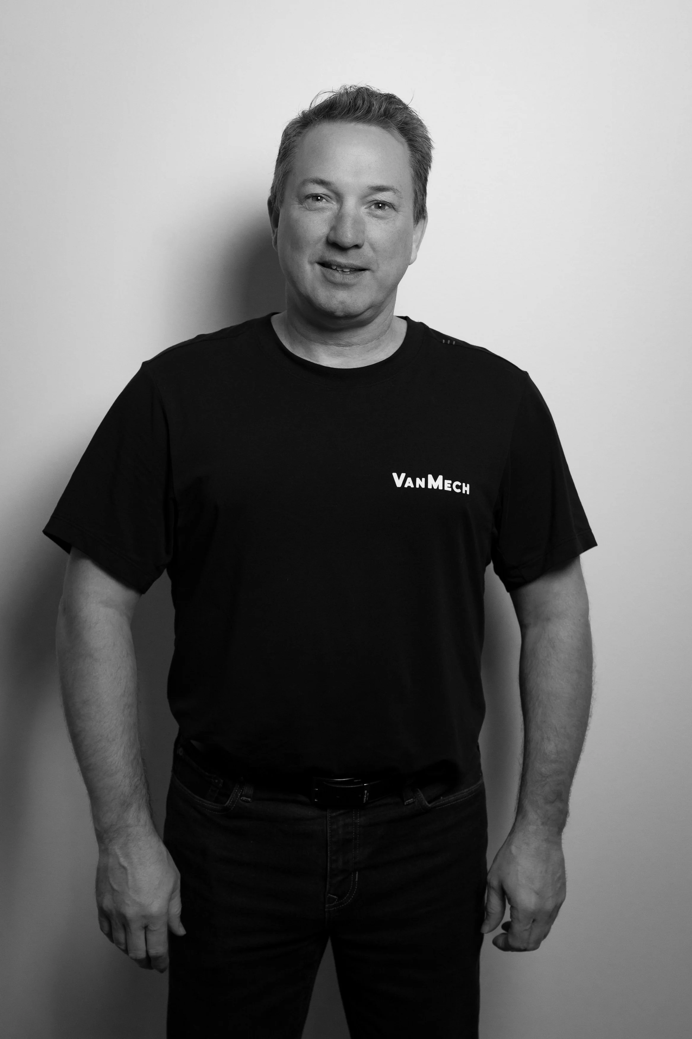 Black and white photo of a man wearing a t-shirt with the word "VanMech" on it, standing against a plain wall.