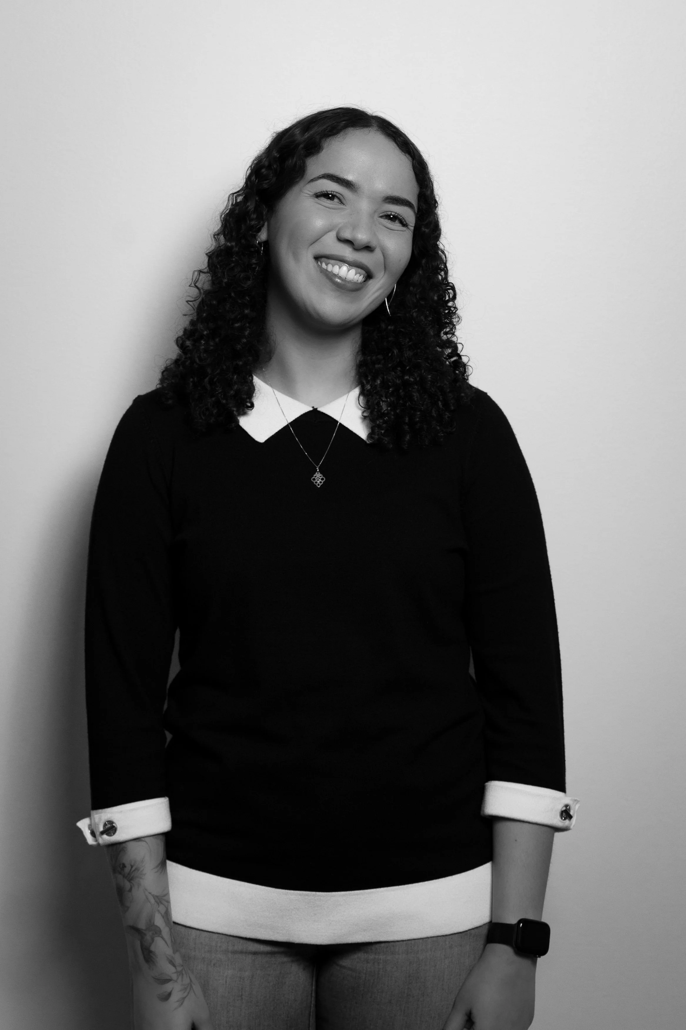 A young woman with curly hair smiling, wearing a long sleeve black shirt with white cuffs, gray pants, a necklace, and an Apple Watch, standing against a plain wall.