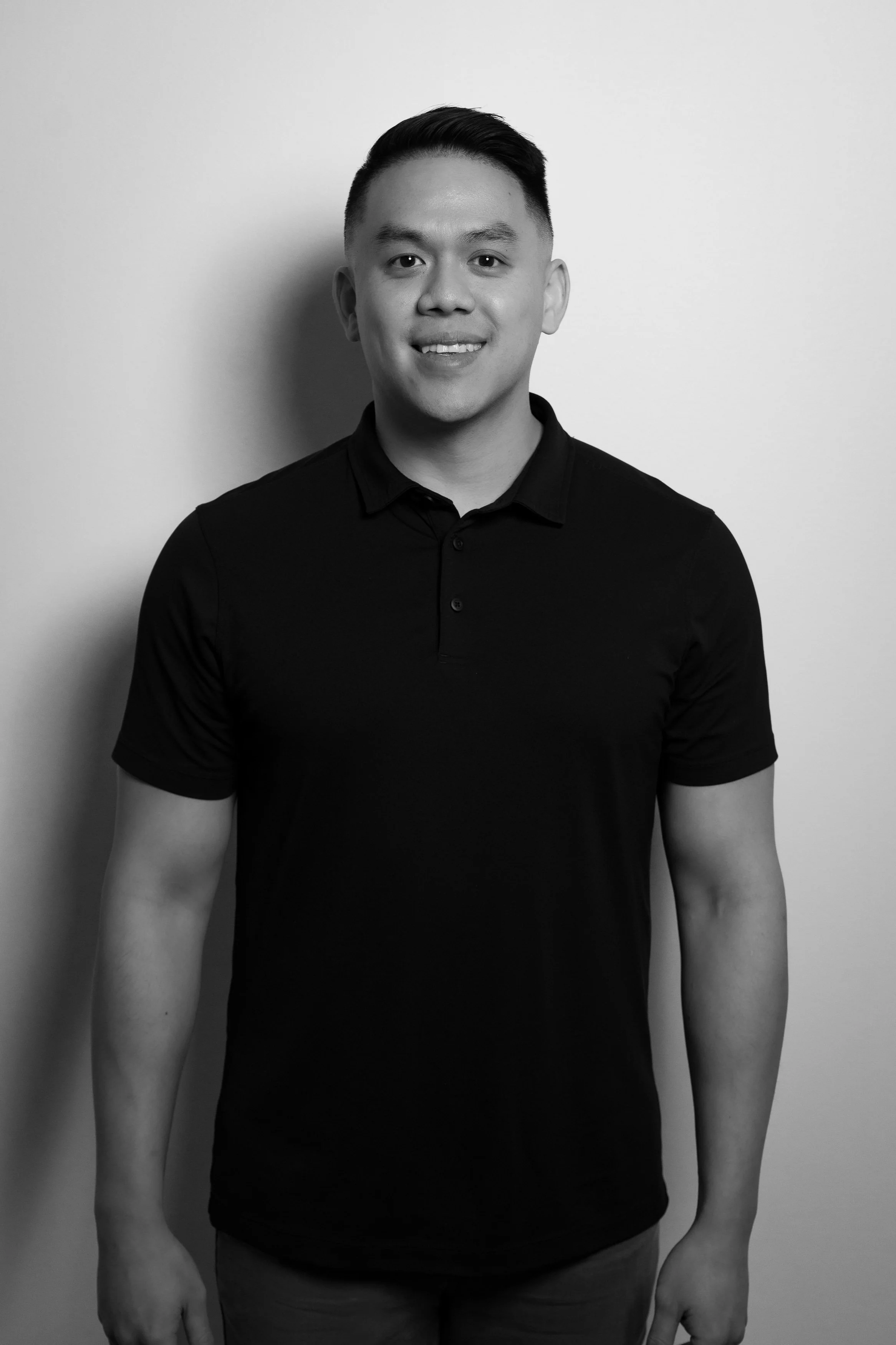 Black and white portrait of a young man with short, dark hair, wearing a short-sleeved, collared shirt, standing against a plain wall, smiling gently.
