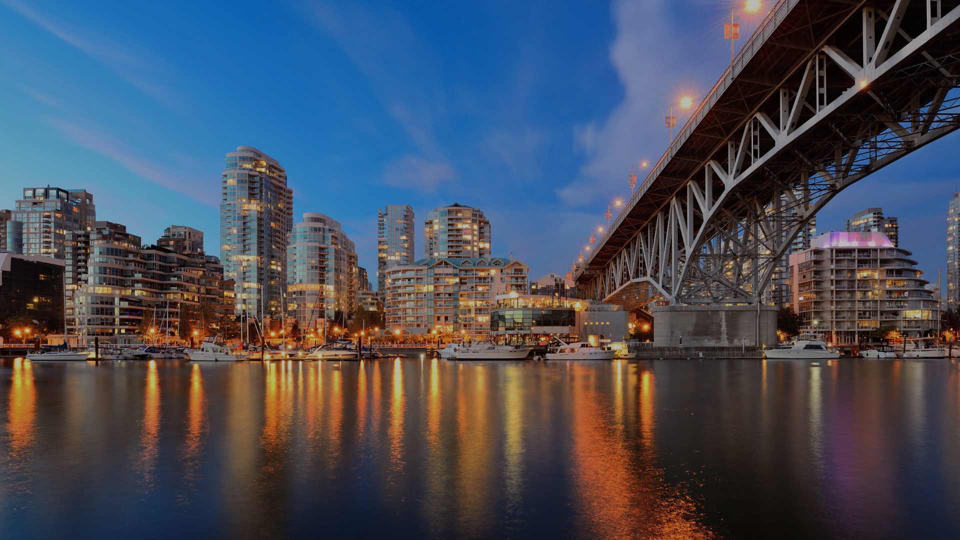 City skyline at dusk with high-rise buildings, marina with boats, and a bridge reflecting lights on the water.