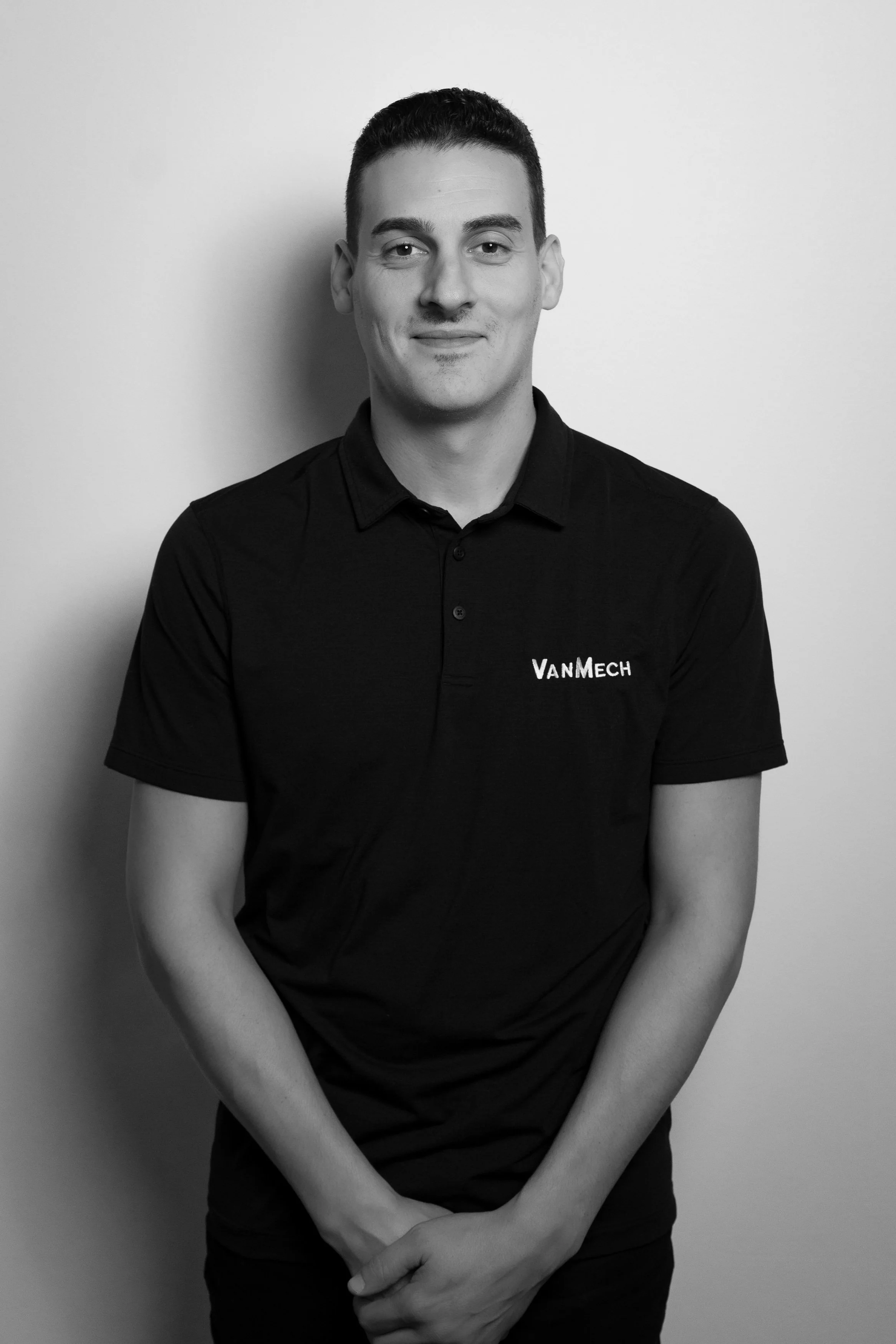 Black and white portrait of a young man with short hair, smiling slightly, wearing a polo shirt with "VanMech" embroidered on it, standing against a plain background.