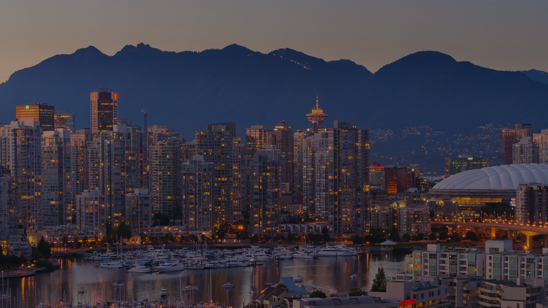 Evening cityscape of Vancouver with tall buildings, mountains in the background, and marina with boats in the foreground.