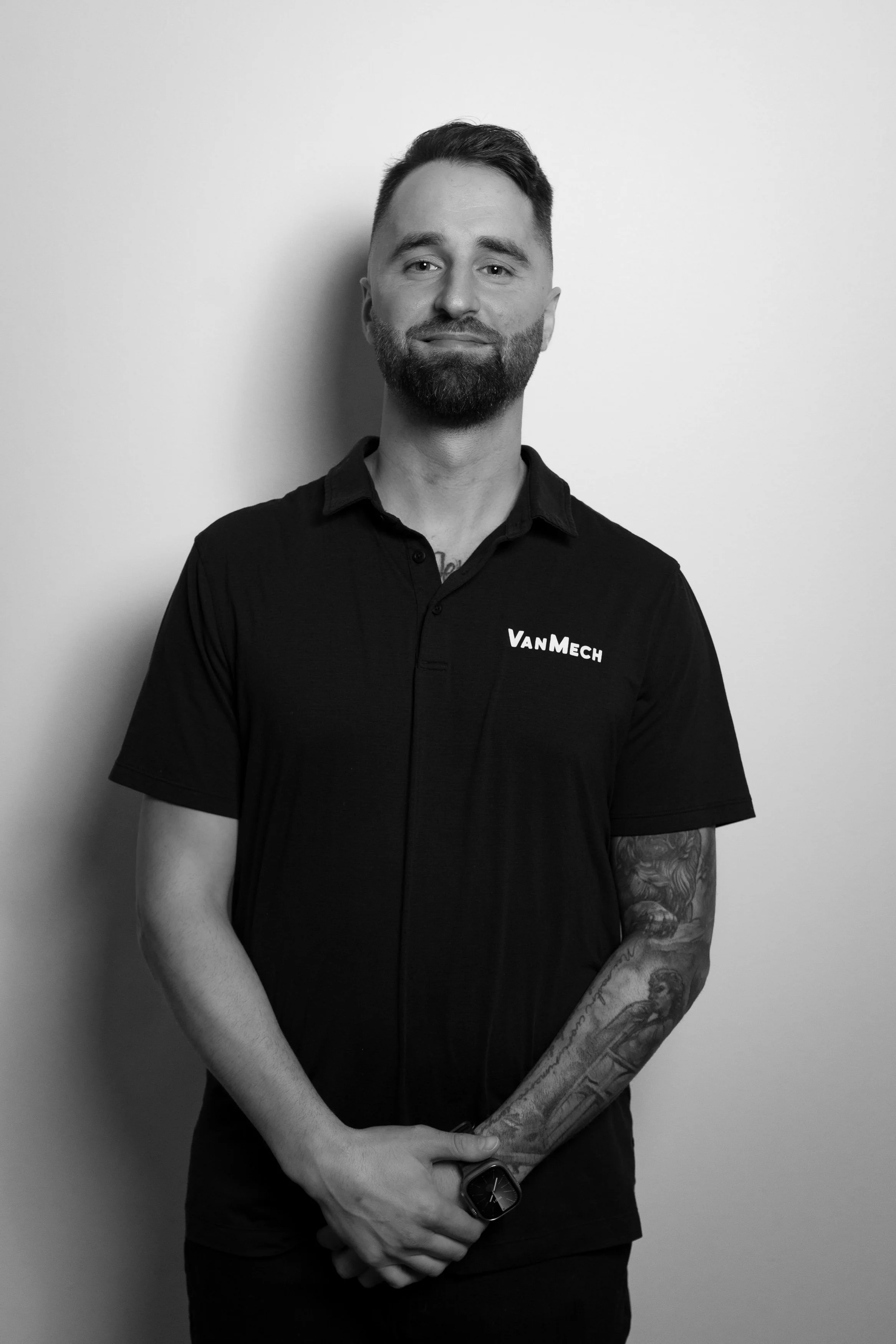 Black and white portrait of a man with a beard and tattoos on his left arm, wearing a polo shirt with 'VanMech' logo, standing against a plain wall.