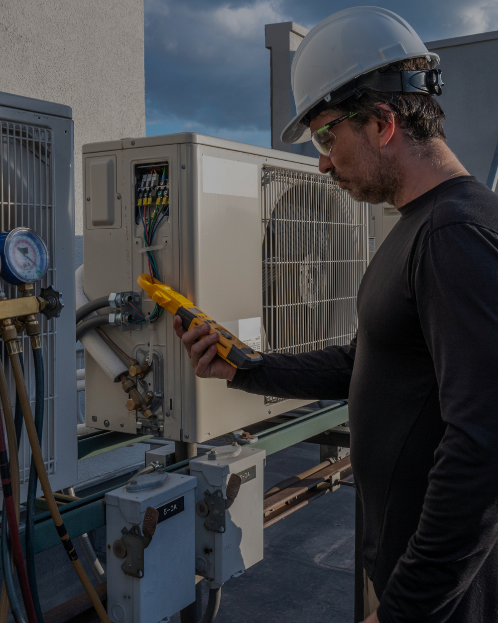 An electrician wearing a white safety helmet and safety glasses uses a multimeter to test HVAC equipment on a rooftop.