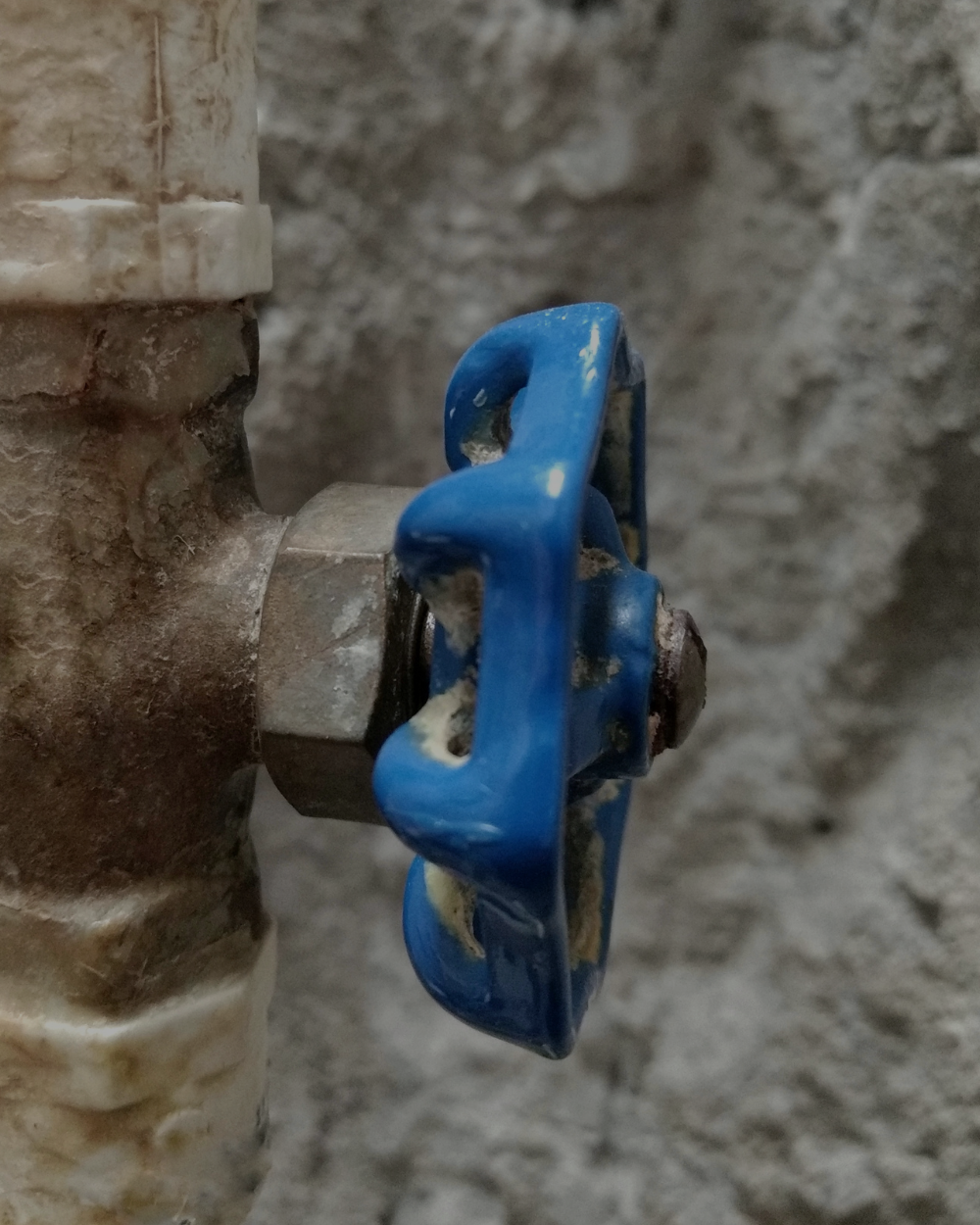 Close-up of a plumbing valve with a blue handle attached to a metal pipe against a rough, gray wall.