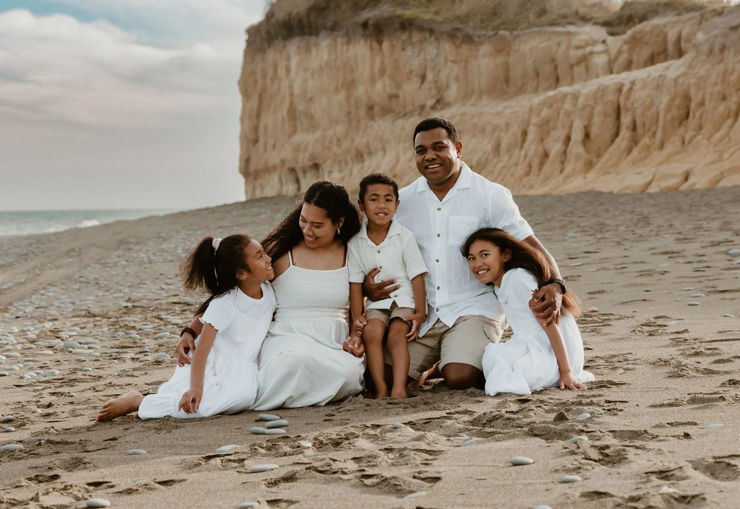 A family of six with three children on a sandy beach with cliffs in the background, all in white clothing, smiling and sitting close together.