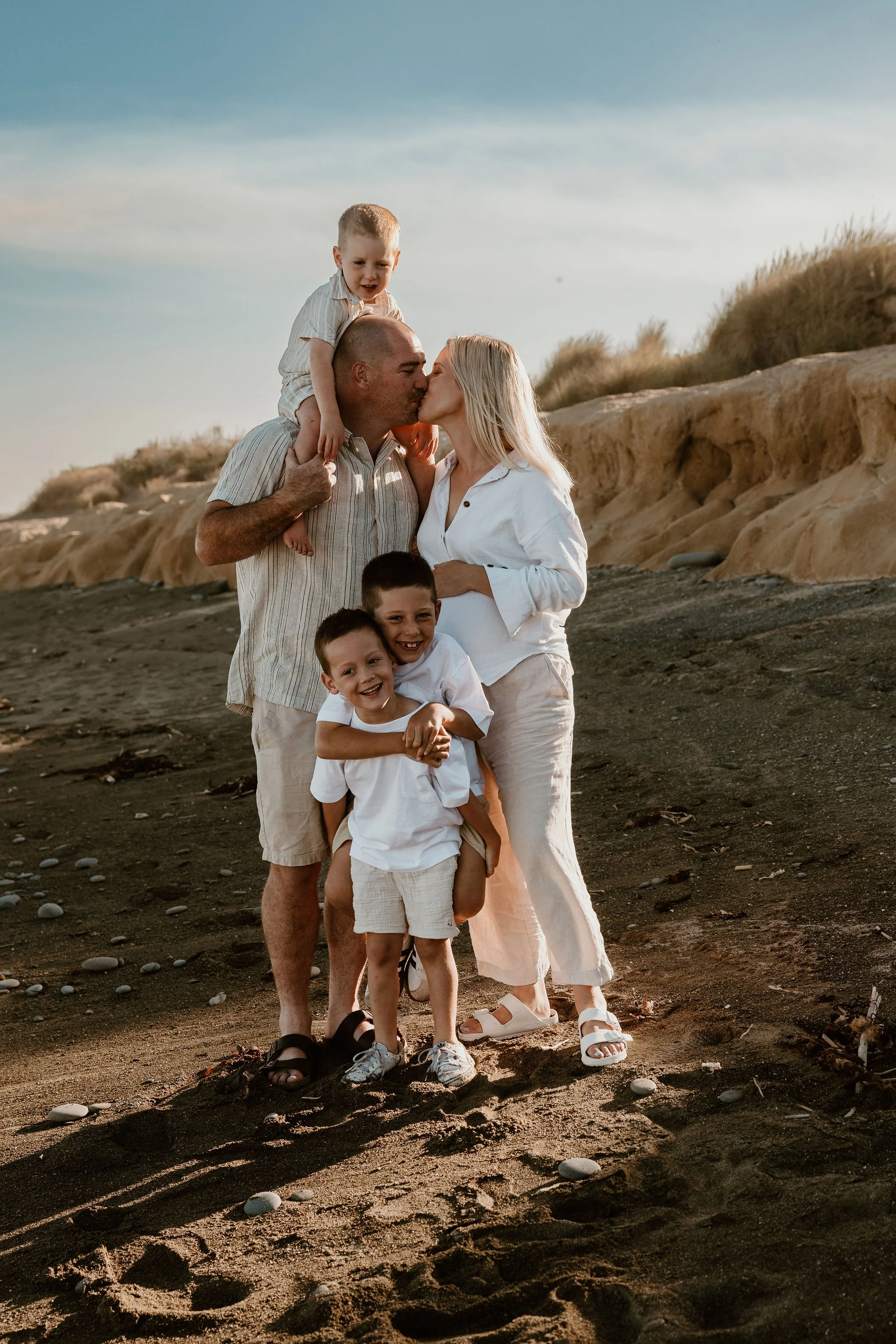 Family of five at the beach, dad and mom kissing, two young boys smiling and one little boy on dad's shoulders