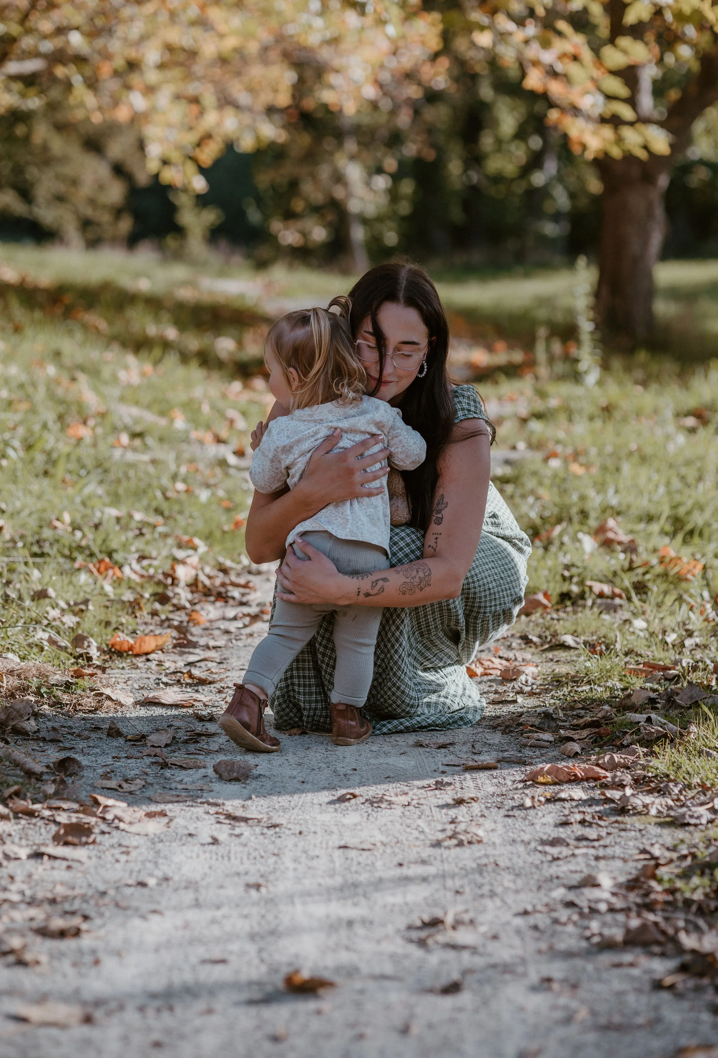 A woman with tattoos and glasses hugging a young girl on a dirt path in a park with fall foliage.