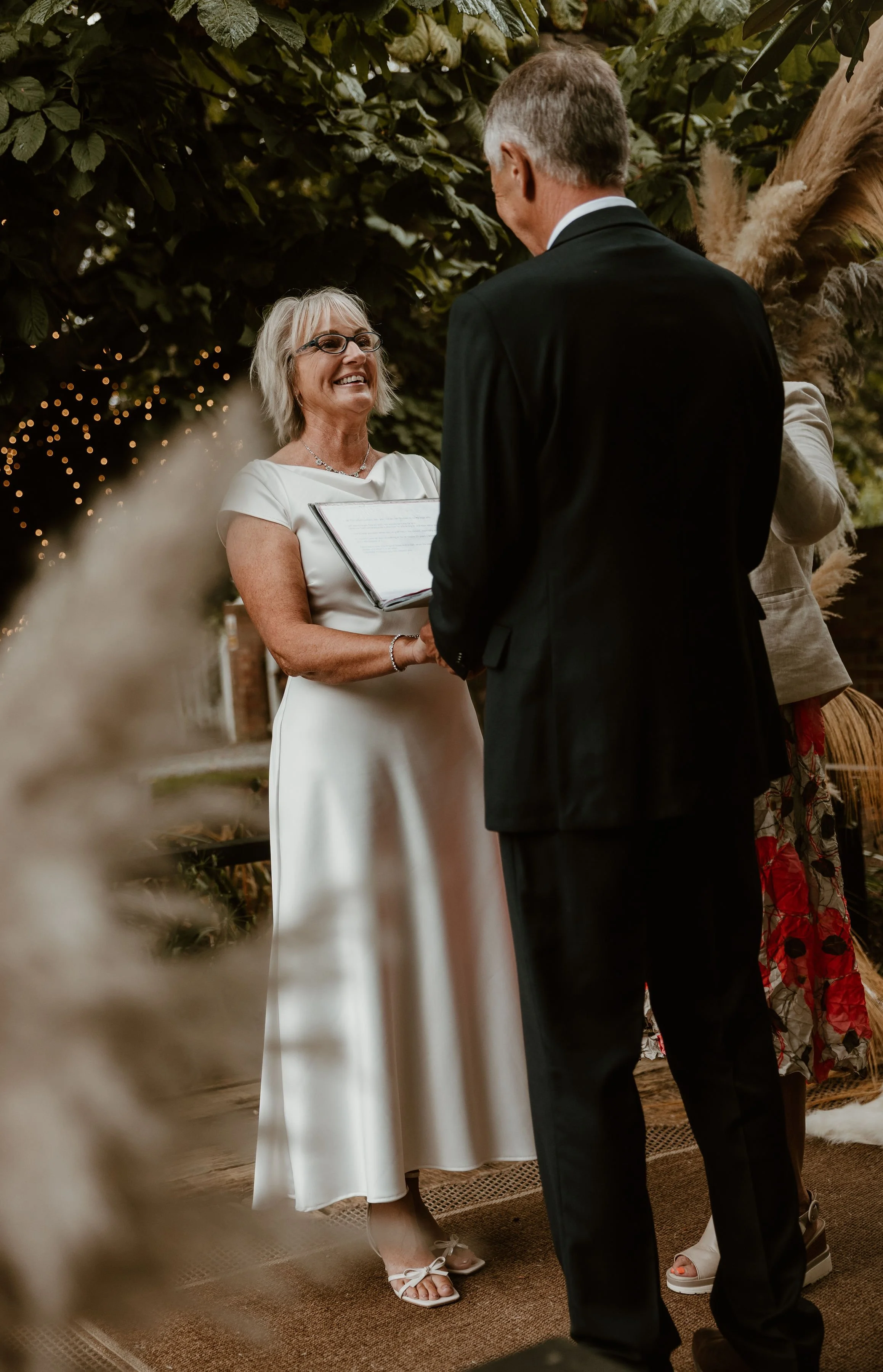 A woman in a white dress reads vows during a wedding ceremony as a man in a black suit listens, under a decorated canopy with greenery and fairy lights.