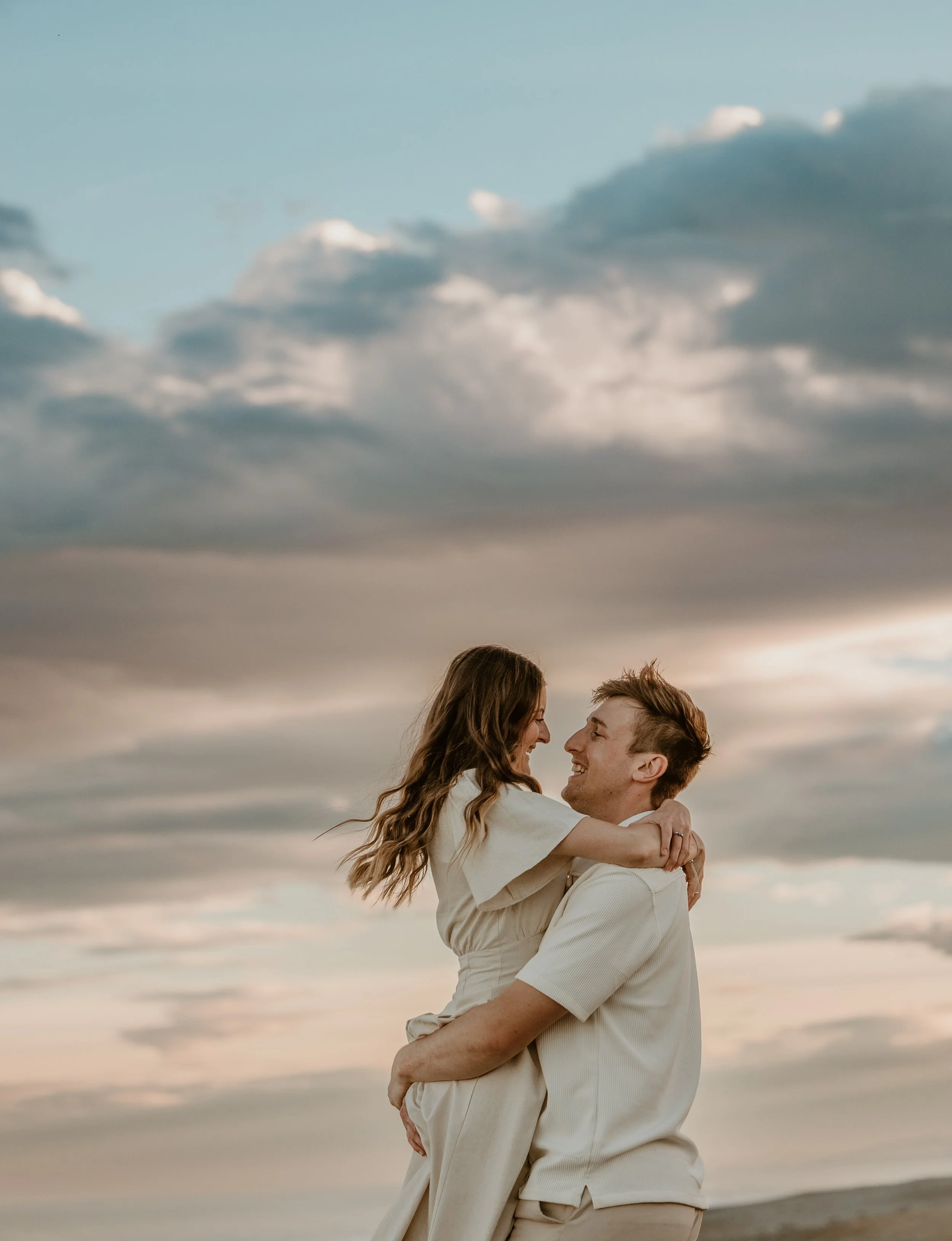 A couple embracing each other outdoors in a field during sunset, with the woman smiling and resting her head on the man's shoulder.