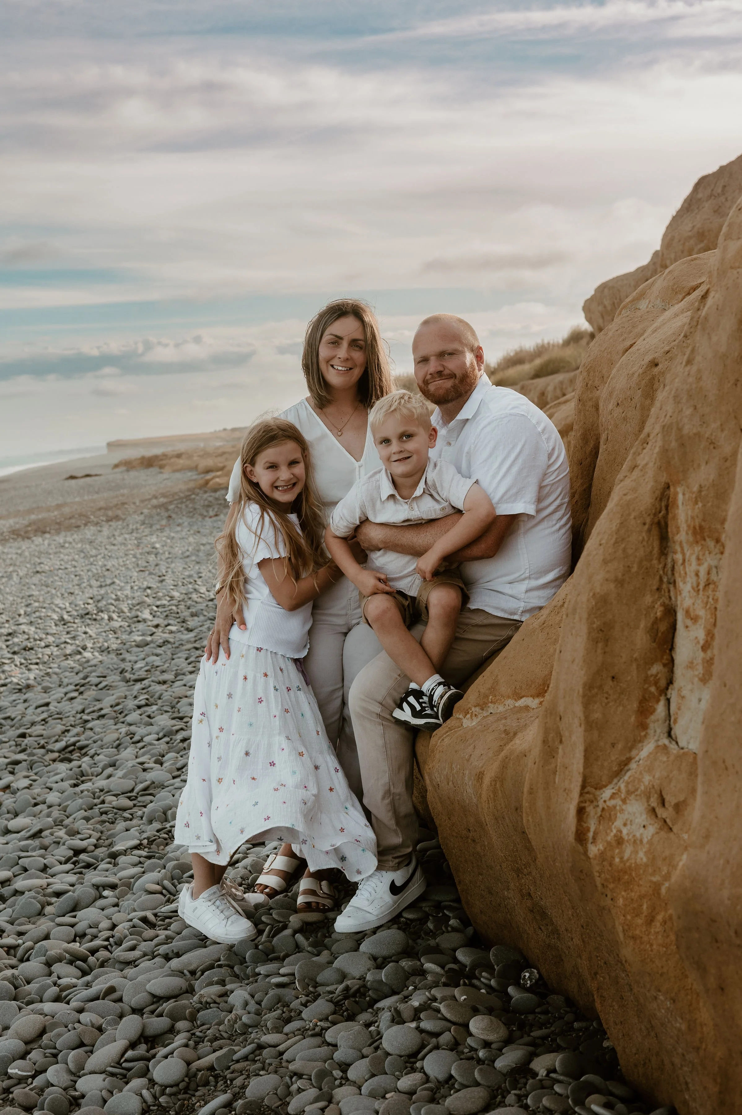 A family of five posing on a pebble beach near rocks with overcast sky in the background.