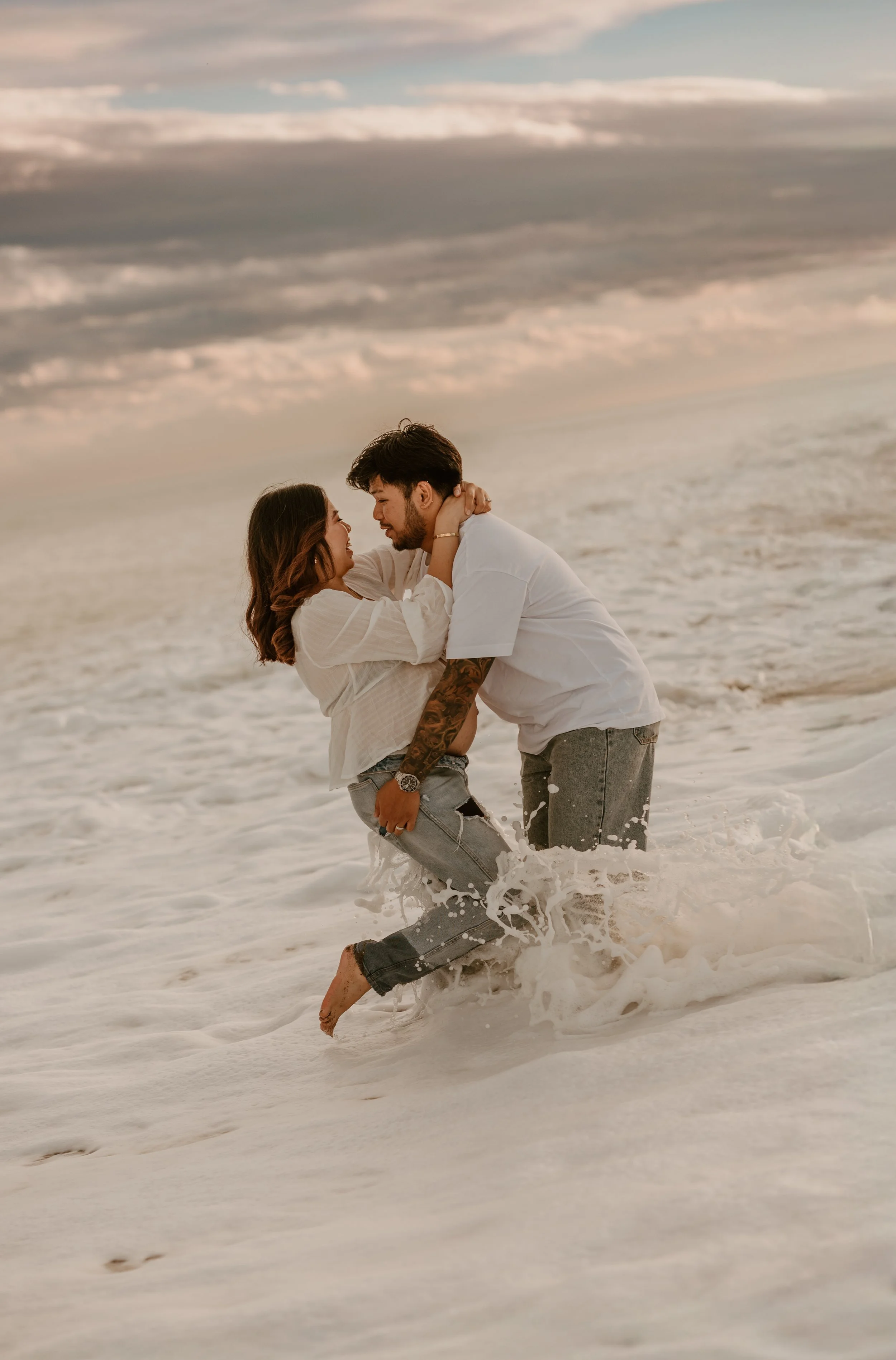 A couple embracing each other outdoors in a field during sunset, with the woman smiling and resting her head on the man's shoulder.