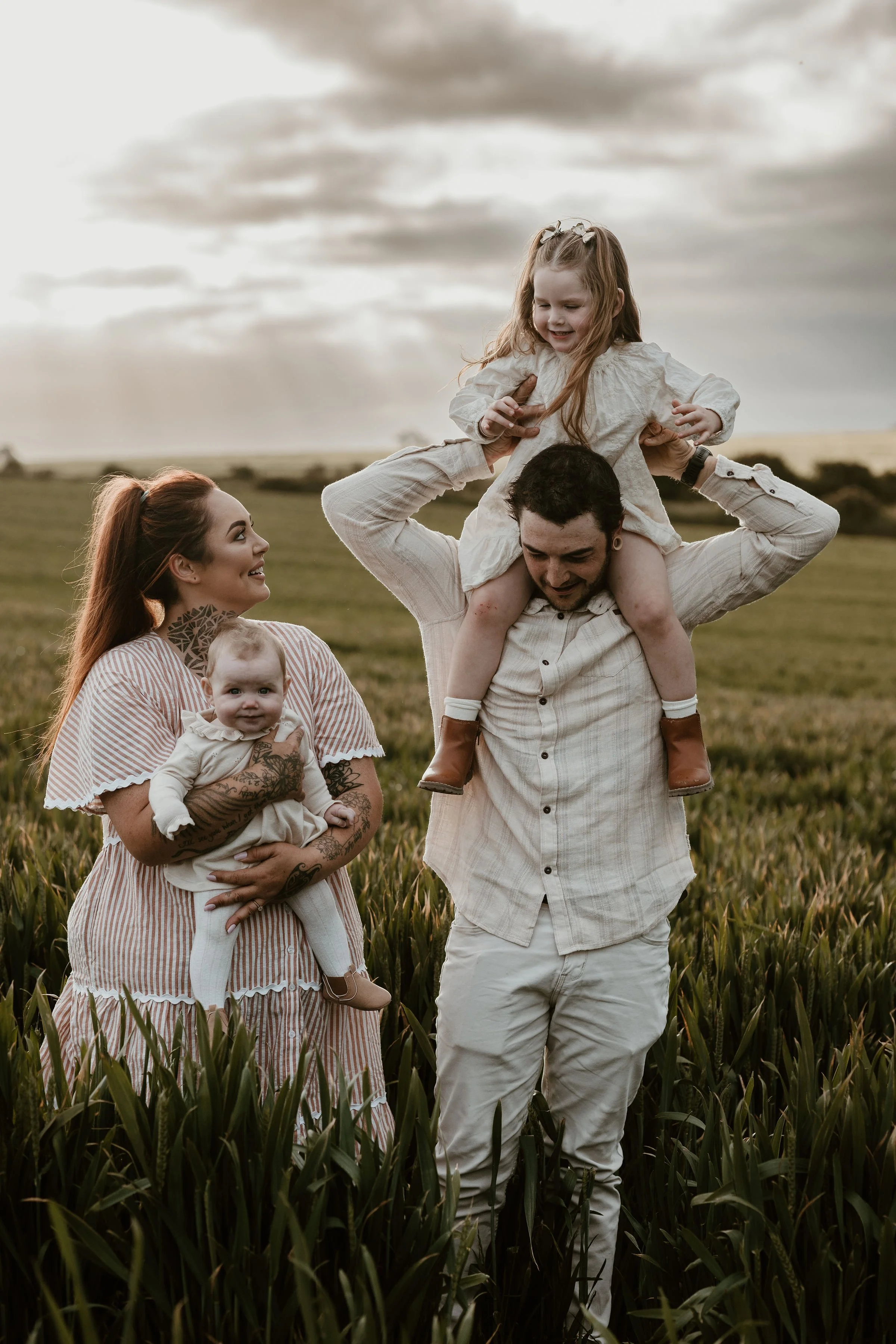 A family of four outdoors in a field during sunset, with a man carrying a young girl on his shoulders, a smiling woman holding a baby girl, all dressed in light-colored clothing.