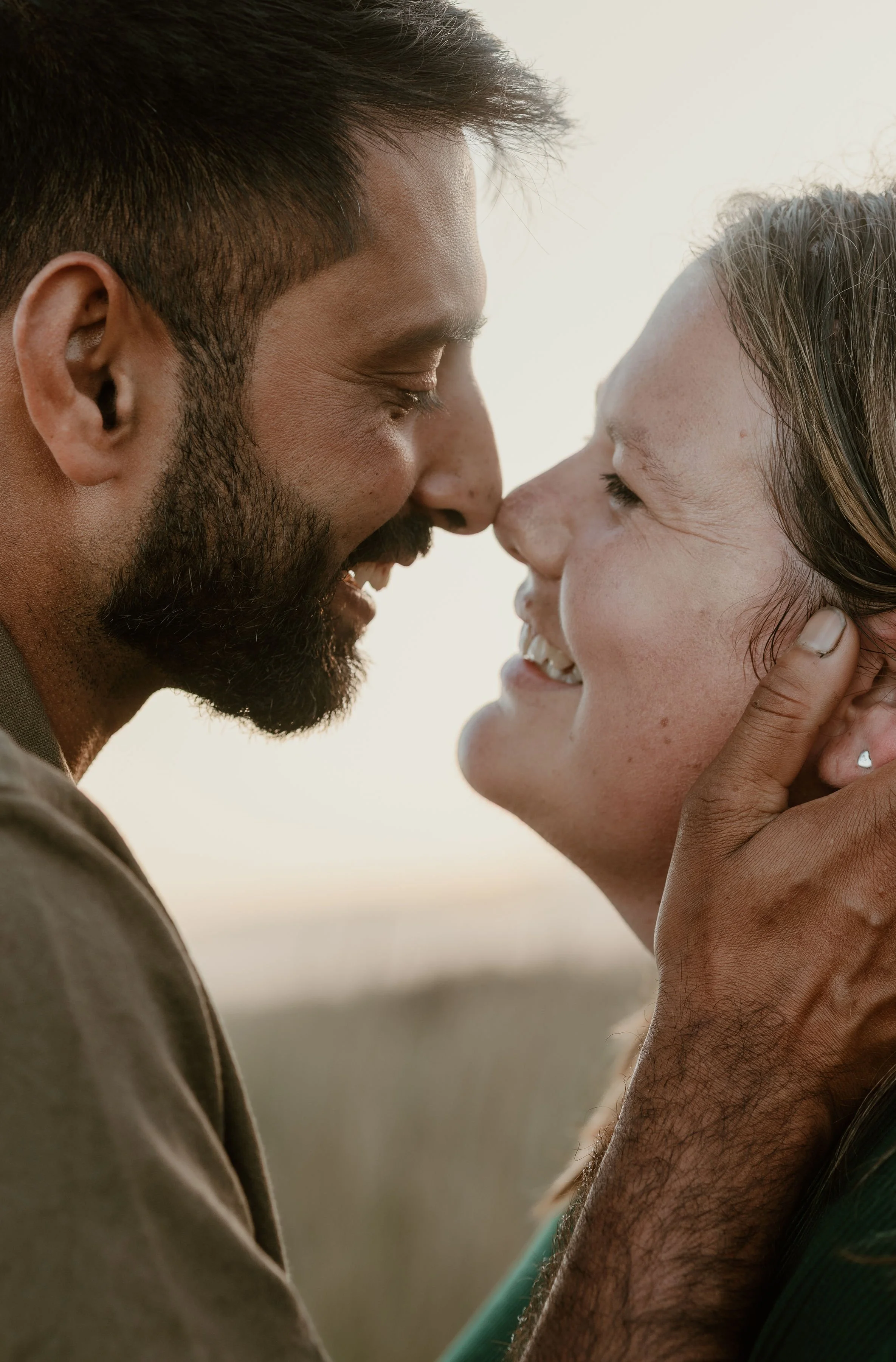 A man and woman with their faces close together, smiling and touching foreheads in an outdoor setting.