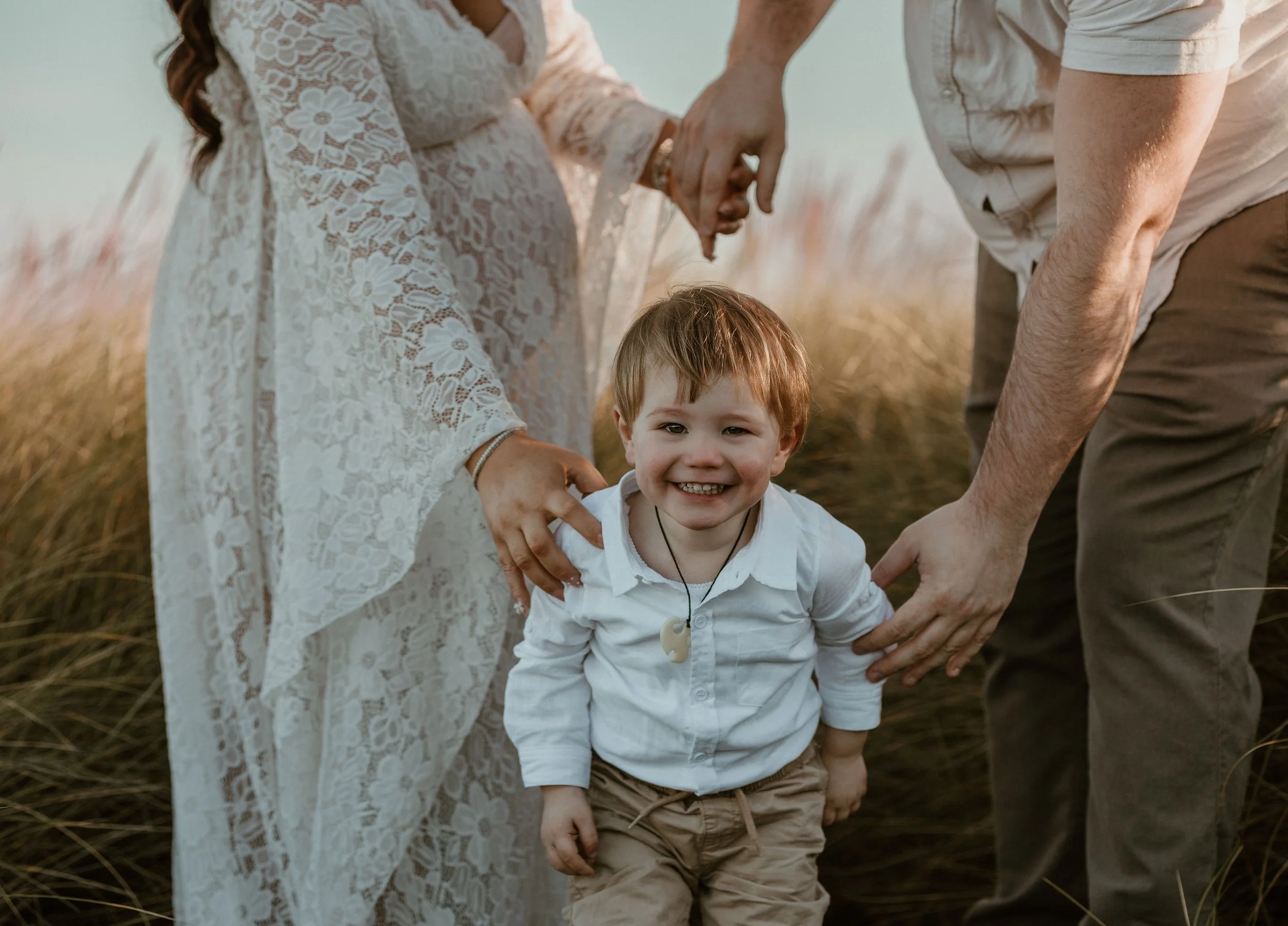 A young boy with a bright smile standing in tall grass, holding hands with a pregnant woman and a man, during a photo shoot outdoors.