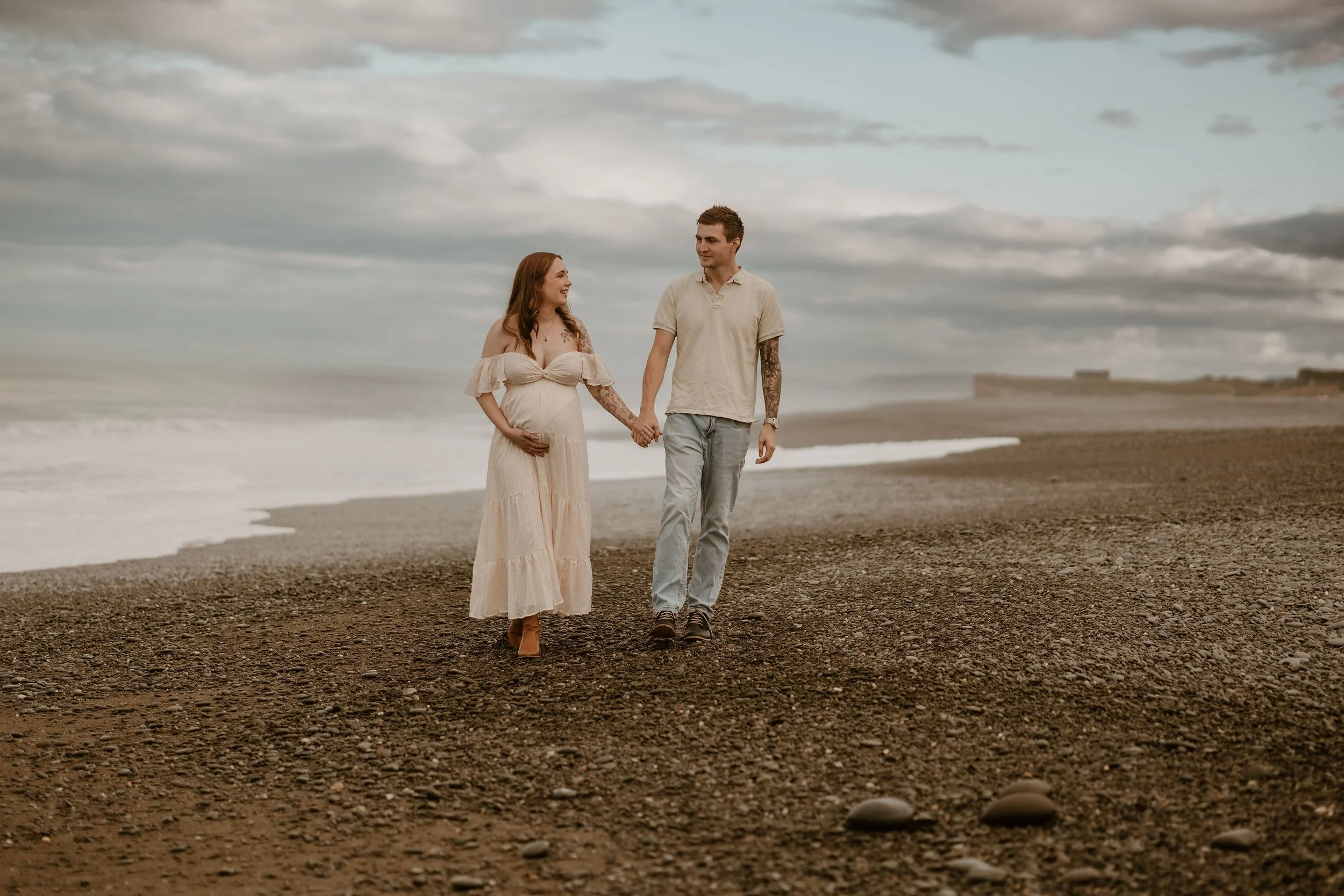 A pregnant woman and a man are walking hand in hand on a pebble beach, with the ocean and cloudy sky in the background.