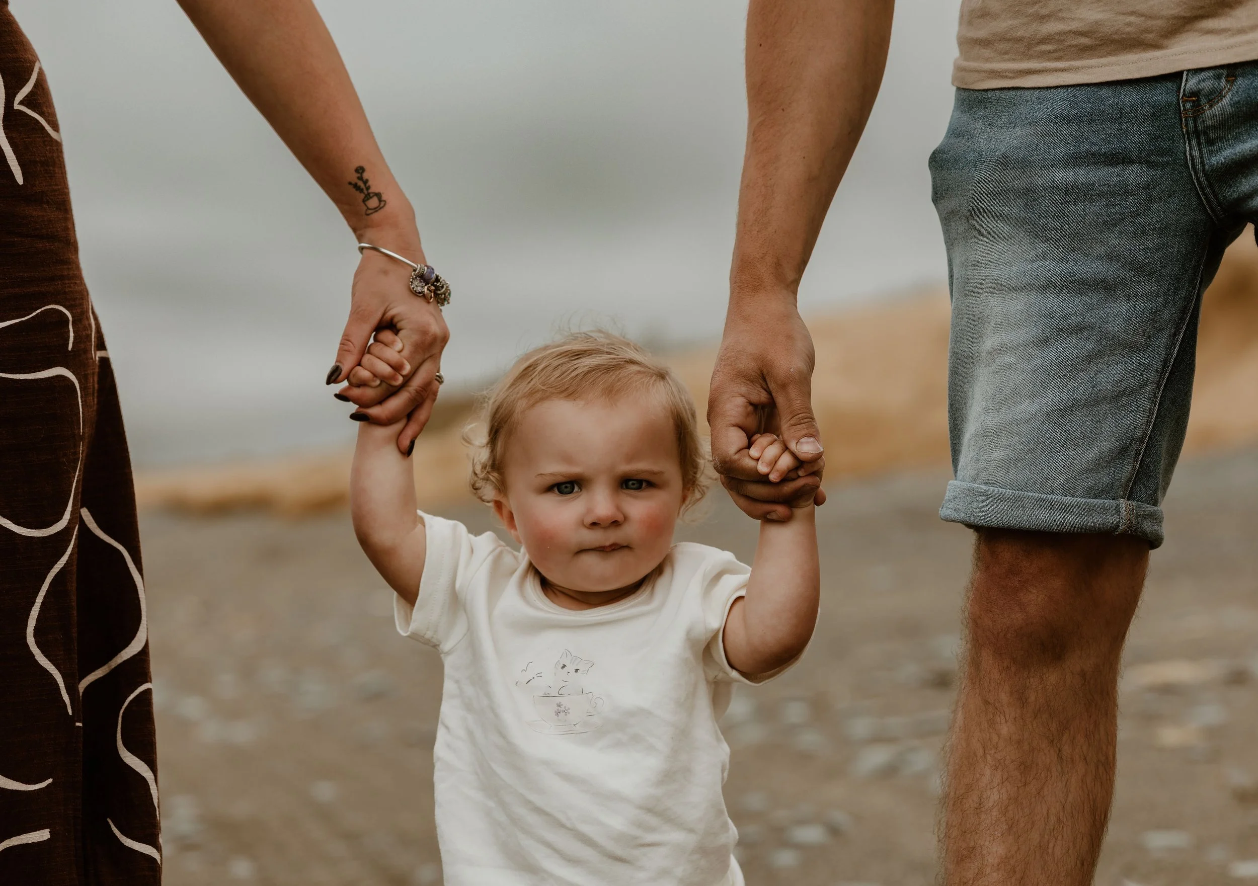 A young child with curly blonde hair and an angry expression, holding onto both parents' hands, standing on a beach with sand and water in the background.