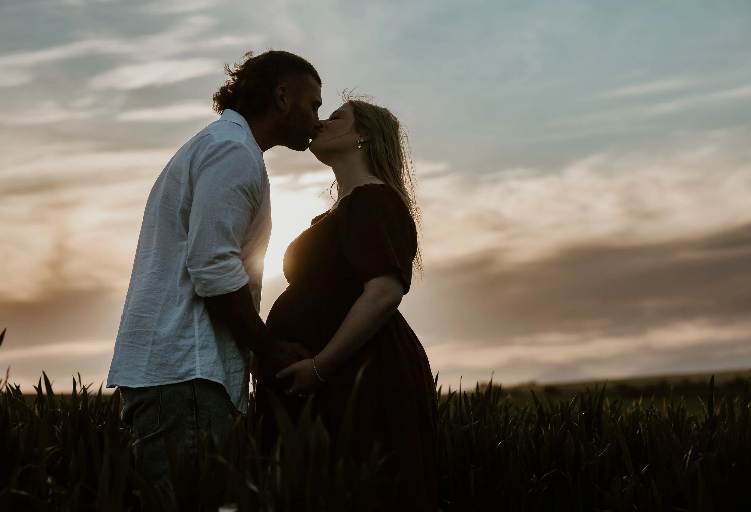 A couple kissing and holding hands in a field during sunset, with a cloudy sky in the background.