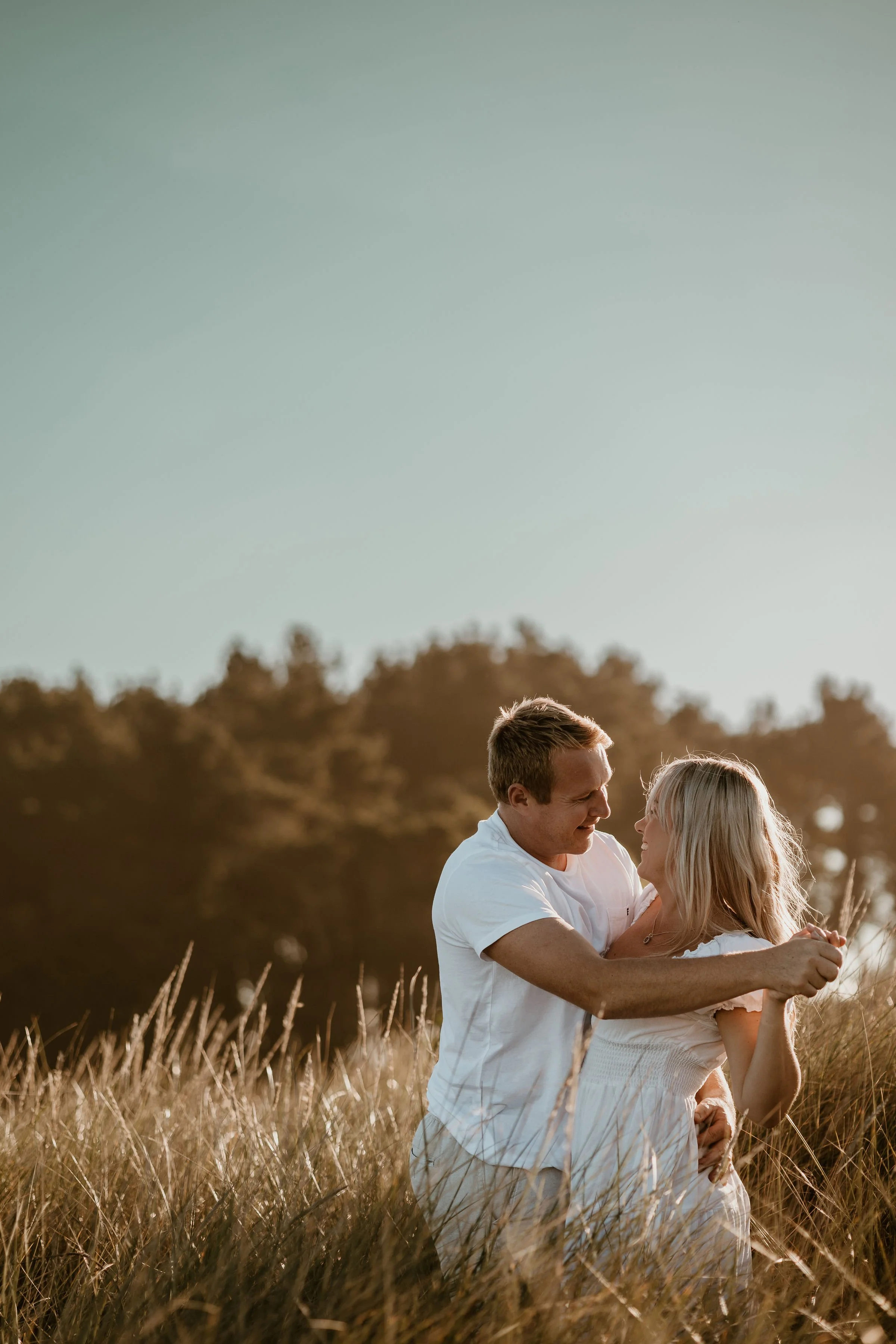 A couple in a field of tall grass during sunset, embracing and looking at each other affectionately.