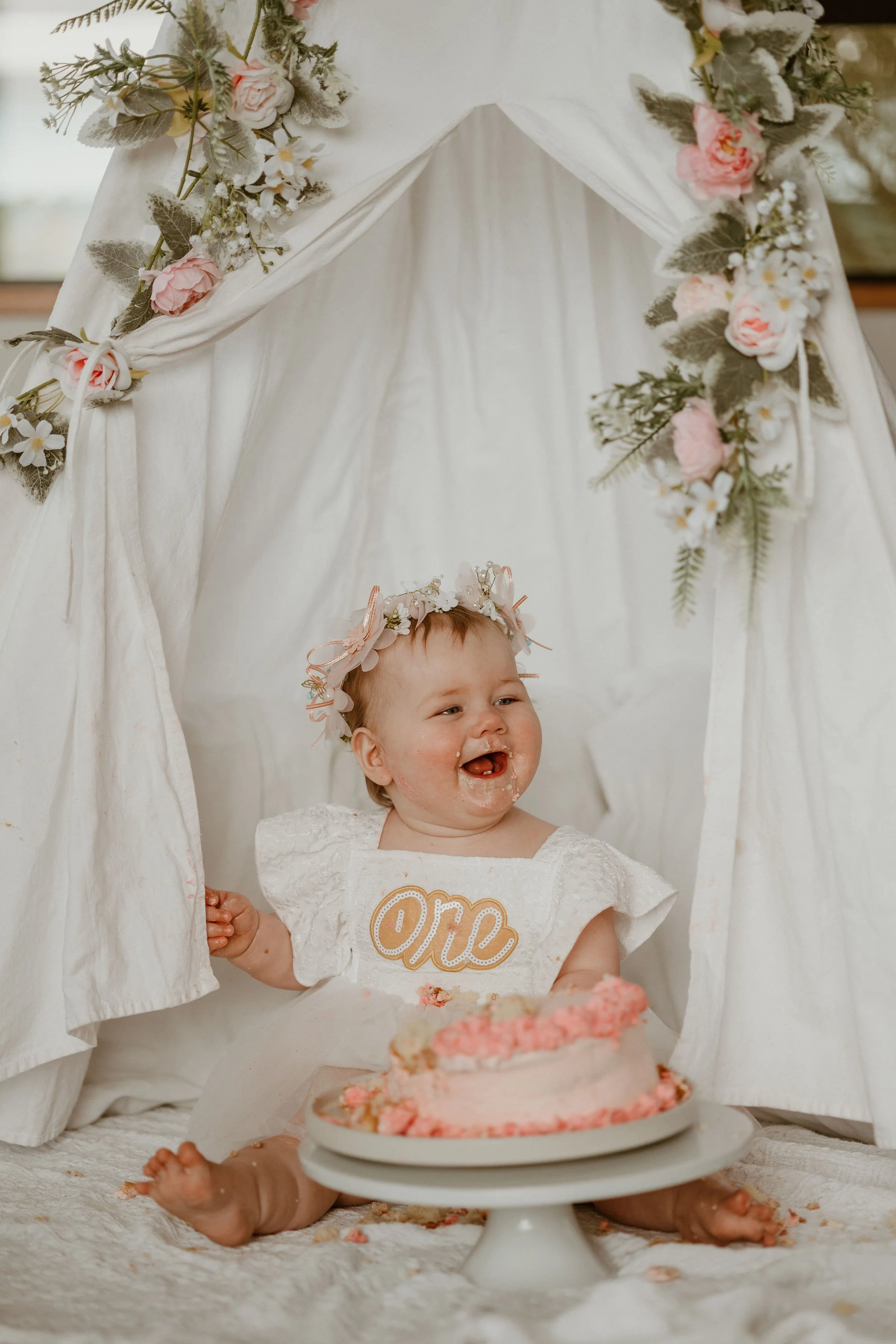 A smiling young child with a floral crown sits inside a white fabric teepee decorated with pink and white flowers, celebrating their first birthday with a pink cake.