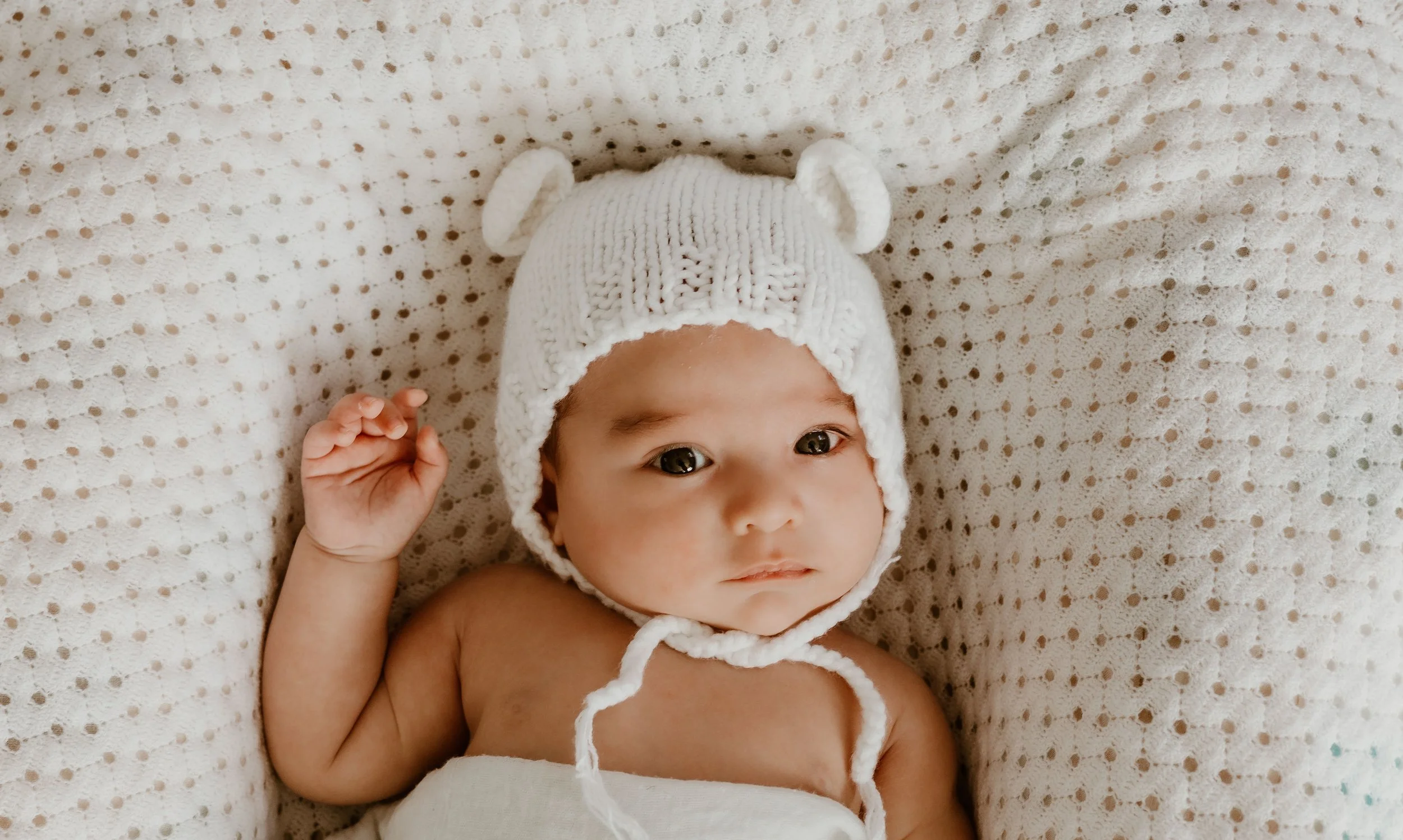A baby lying on a knitted blanket, wearing a white knit hat with bear ears.