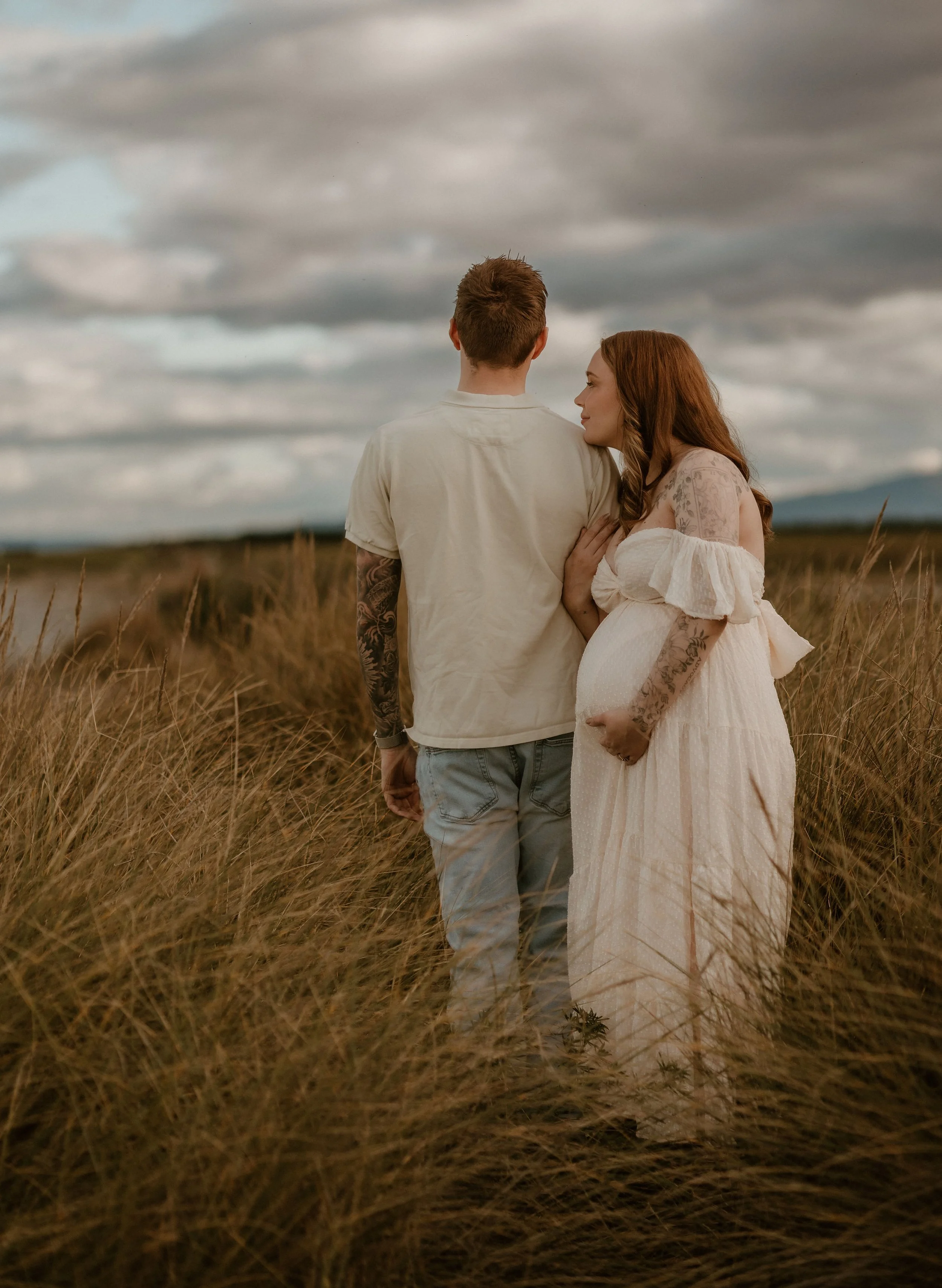 A pregnant woman and a man standing in a grassy field under a cloudy sky, with the woman gently touching her belly and the man looking away.