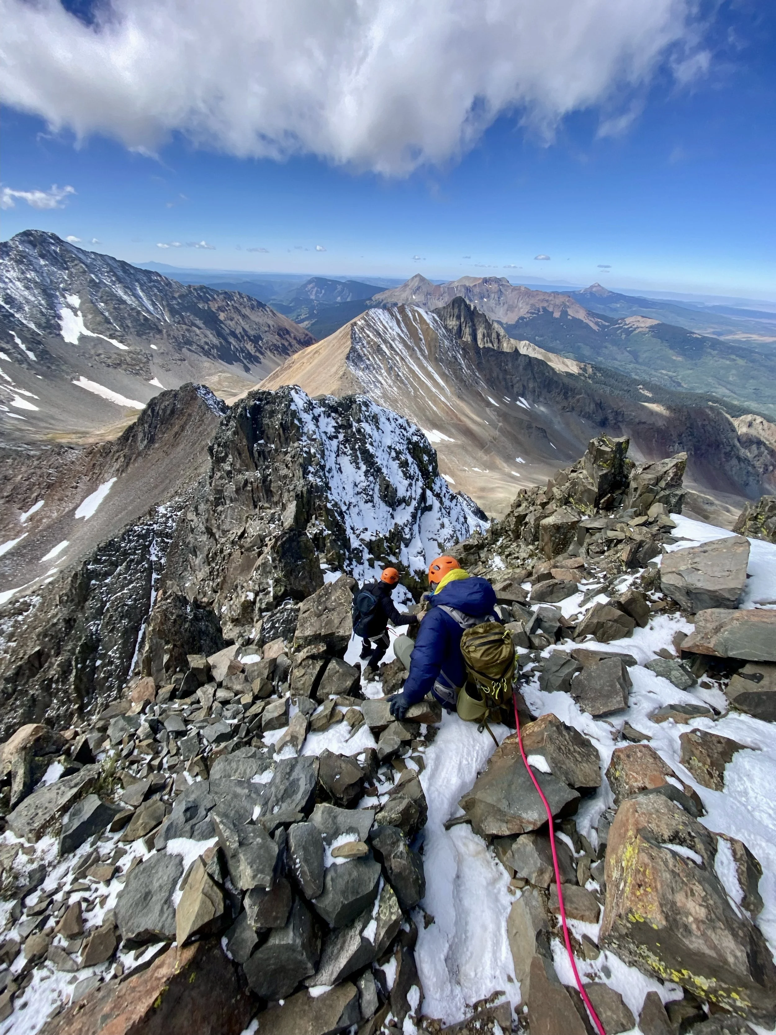 Three climbers with helmets and backpacks navigating a rocky, snow-dusted mountain ridge with a vast mountain range and blue sky in the background.