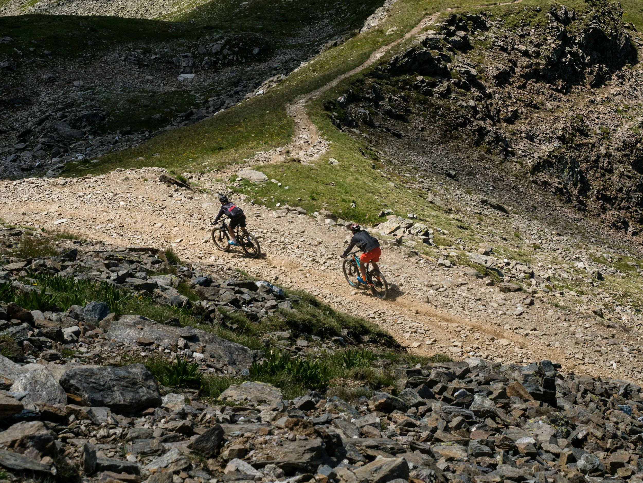 Two mountain bikers riding along a rocky trail on a steep hillside with grass and mountain rocks.
