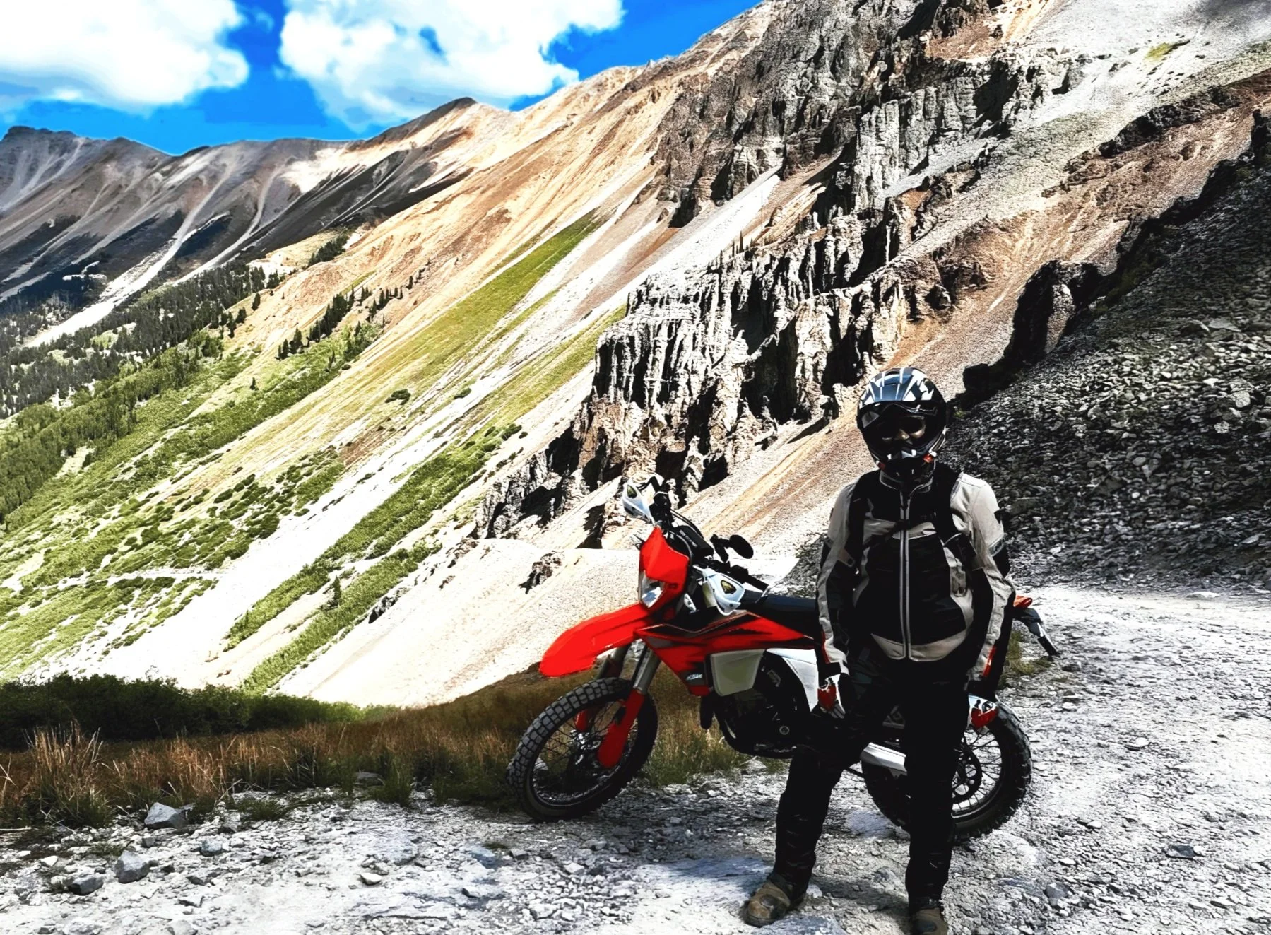 A motorcyclist in adventure gear standing next to a red and black off-road motorcycle on a mountain trail with steep, rocky slopes and green vegetation, under a partly cloudy sky.