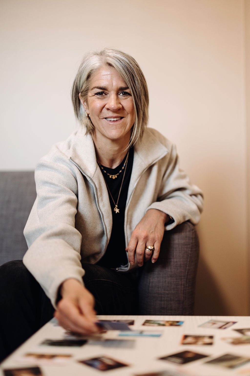 Woman with short gray hair smiling, wearing a beige hoodie and necklaces, sitting on a gray couch, reaching for photos on a table.