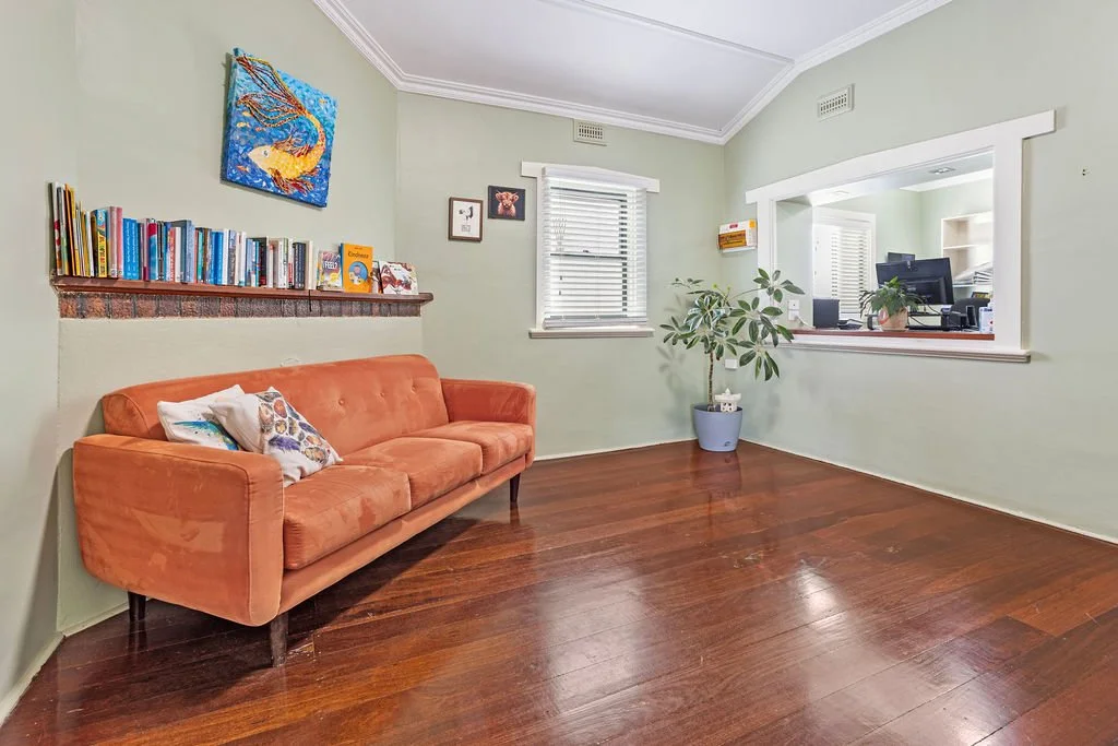 Living room with green walls, an orange sofa with pillows, a bookshelf with books, a window with blinds, a potted plant, and a pass-through window opening to a home office area.
