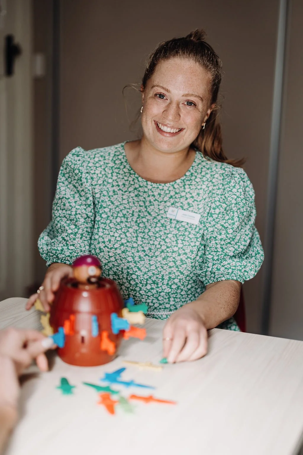 A smiling woman with red hair tied back, wearing a green floral dress, sitting at a table playing a board game with colorful pieces and a Pokémon-themed spinner.