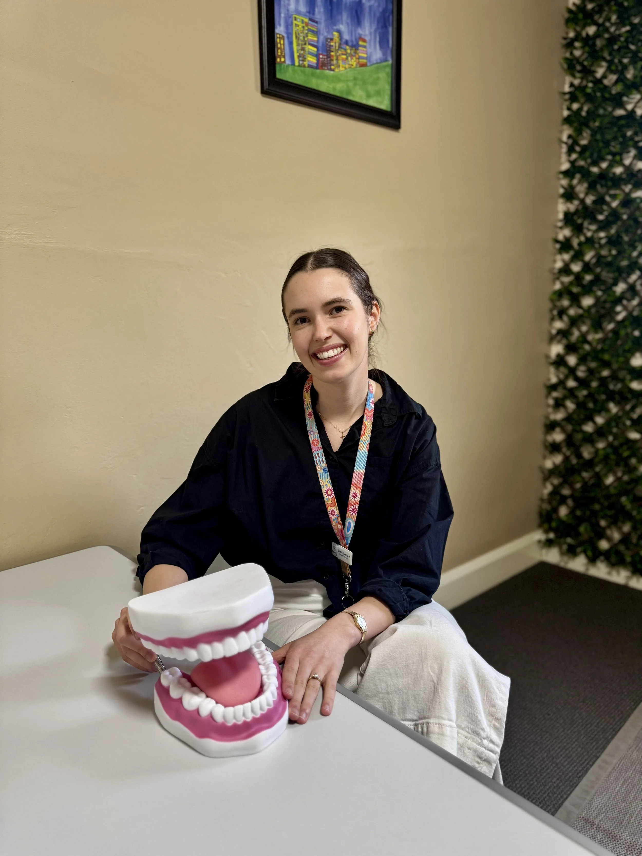 A young woman with dark hair, wearing a black shirt and beige pants, sitting at a table with a large mouth model with teeth and tongue, smiling at the camera. There is a colorful painting on the beige wall behind her and a green plant to the right.