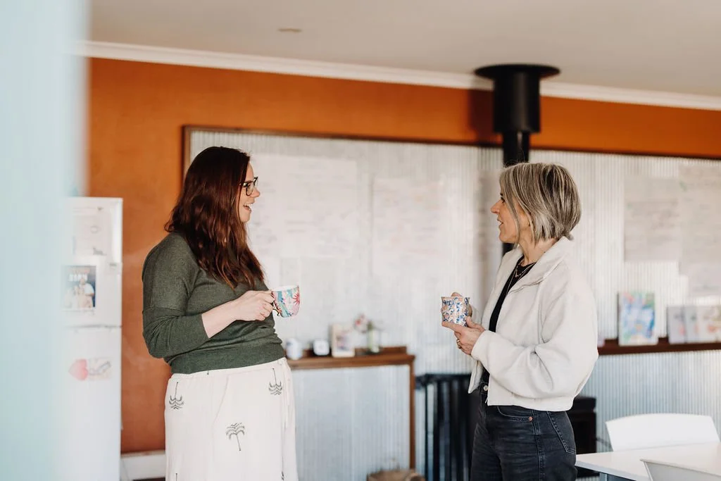 Two women having a conversation, each holding a mug, in a cozy indoor setting with warm colors and artwork on the walls.