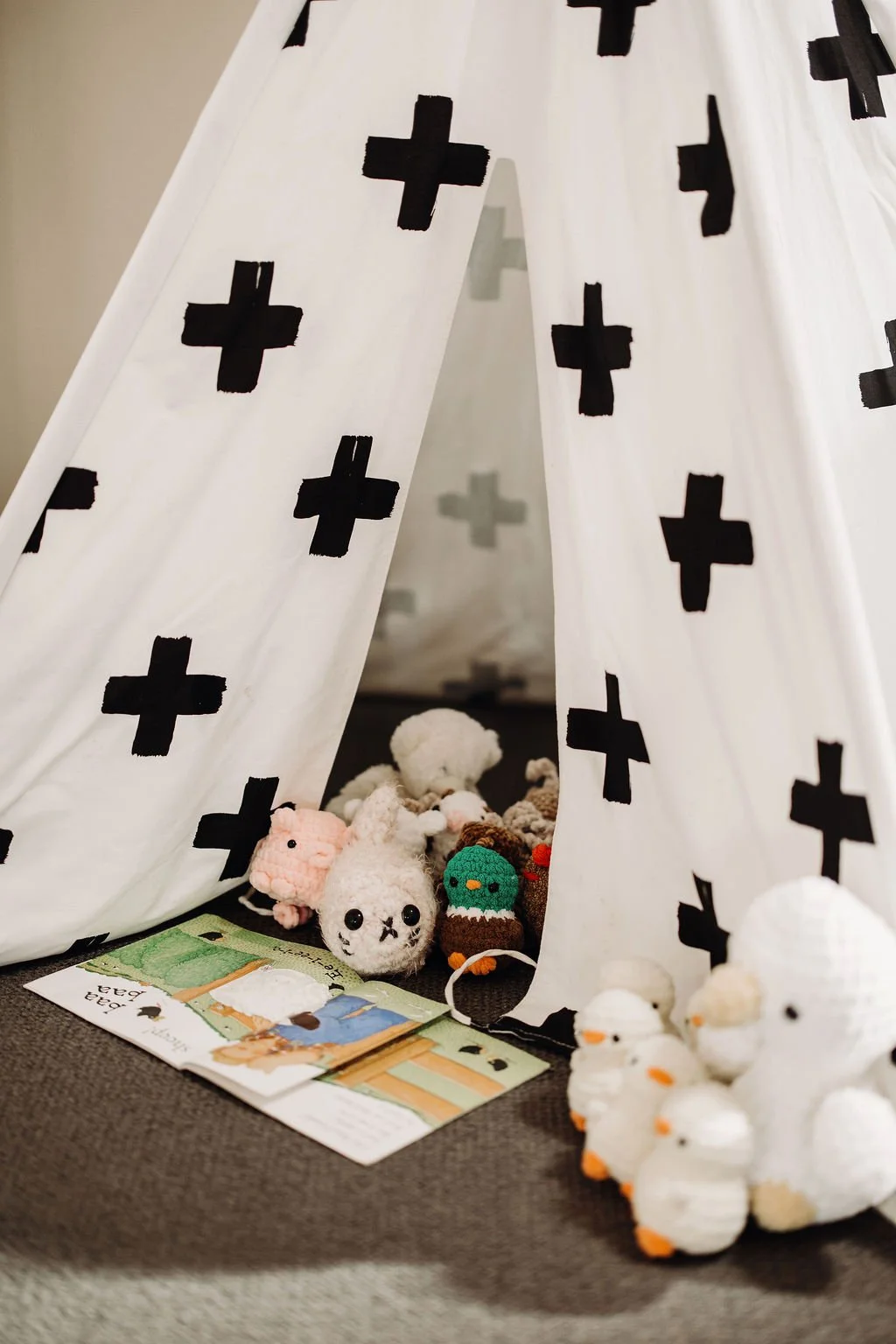 A white teepee with black plus signs, filled with plush toys including animals and a book, on a dark carpeted floor.