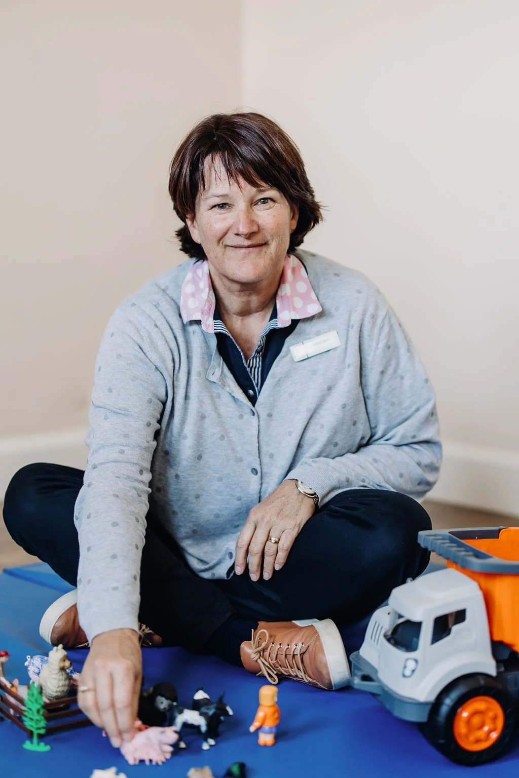 Woman sitting on the floor surrounded by toy farm animals and a toy truck, wearing a gray sweater with pink polka dots and a striped shirt underneath.
