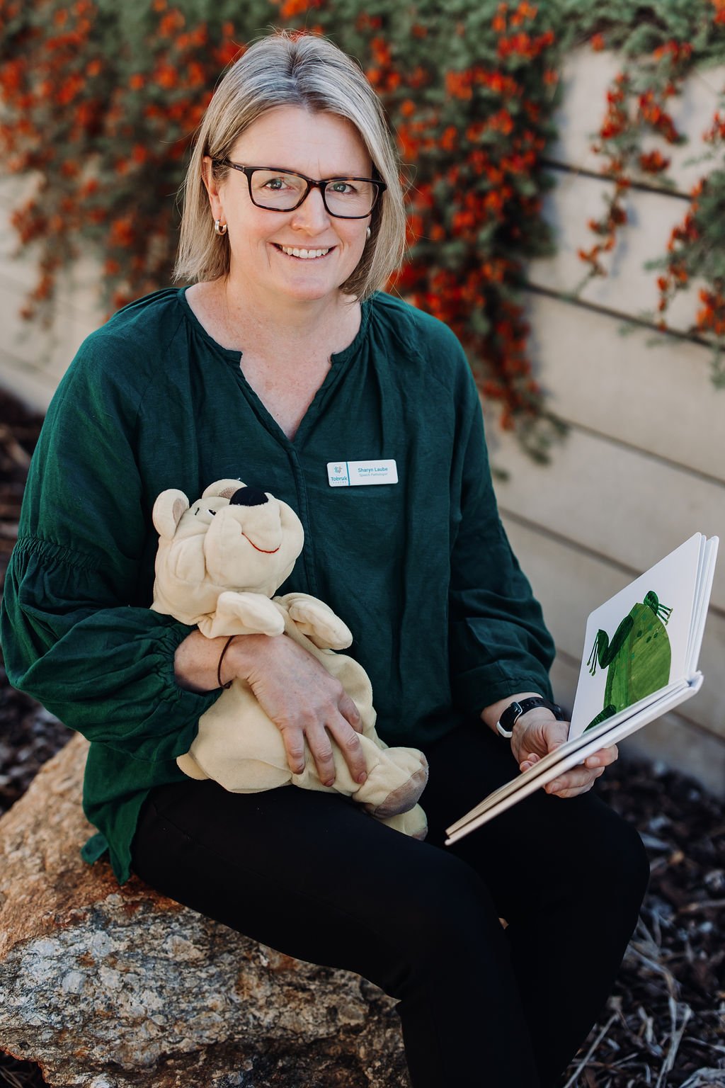 A woman with glasses, wearing a dark green blouse, sitting outdoors on a rock, holding a plush stuffed toy and reading a children's book with a frog illustration. There is a beige wall and red-orange flowers in the background.
