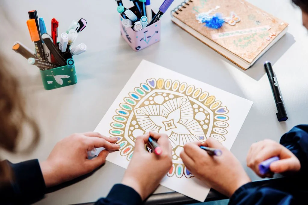 Two people coloring intricate geometric design on white paper with markers, surrounded by pens in decorative cups and other art supplies on a light-colored table.