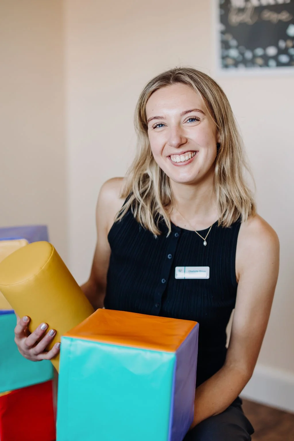 A smiling woman wearing a black sleeveless top and a name tag holding colorful gift boxes in a room.