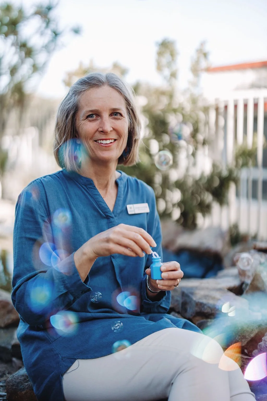 A smiling woman with gray hair, wearing a blue shirt, sitting outdoors and blowing bubbles.