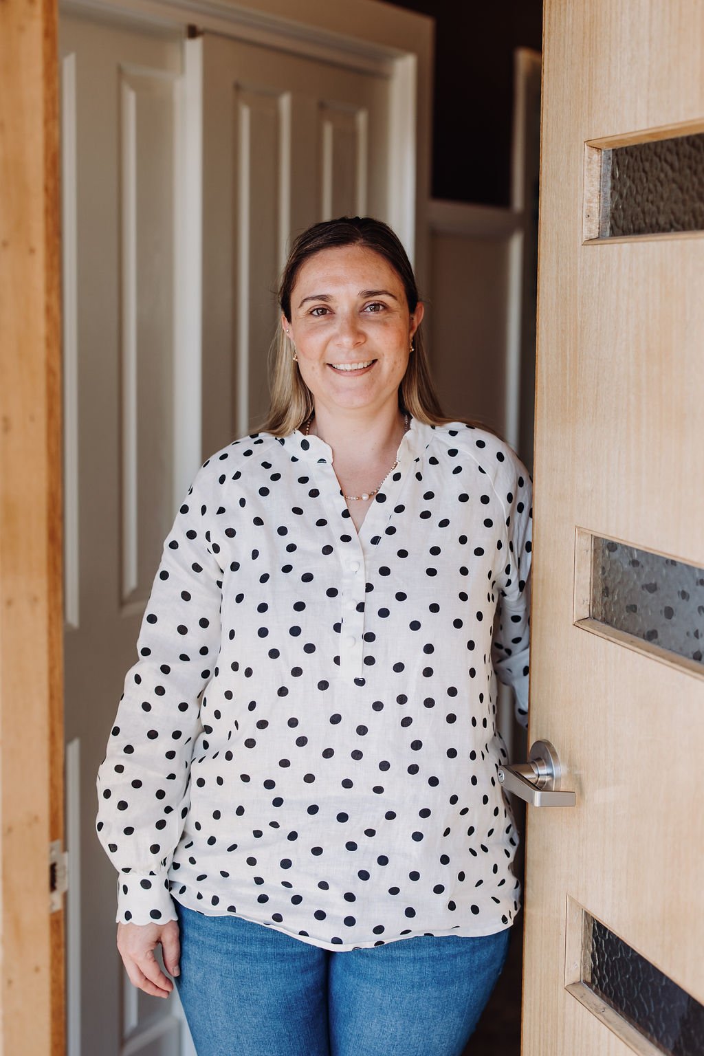 Woman in a white polka dot blouse standing in a doorway, smiling.