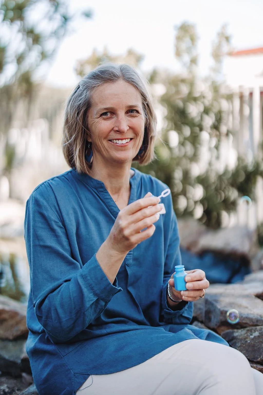 A smiling woman with shoulder-length gray hair, wearing a blue top and earrings, sitting outdoors by a pond or stream, blowing bubbles with a bubble wand.